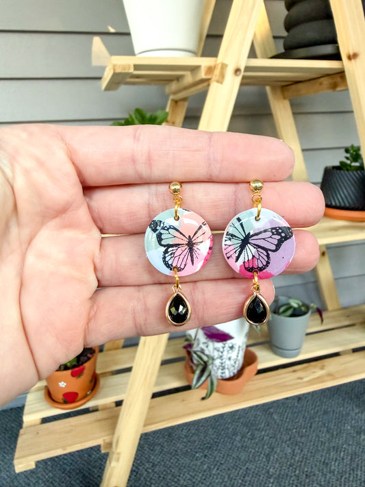 Hand holding colorful earrings with a black butterfly stencil design, and a black charm hanging down. Held against a background with plants. 