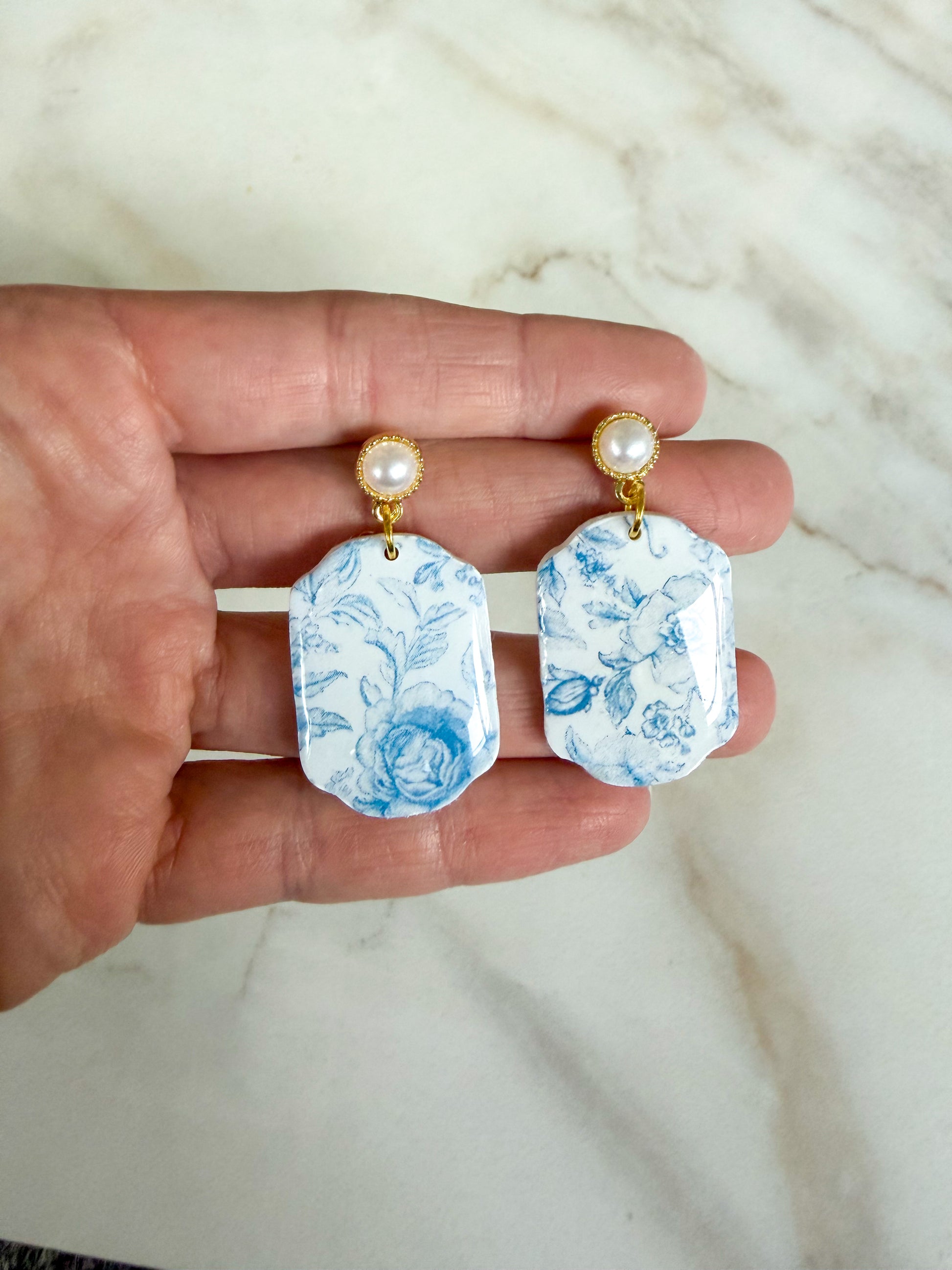 Hand holding a pair of blue floral earrings with pearl-like studs on a marble surface.