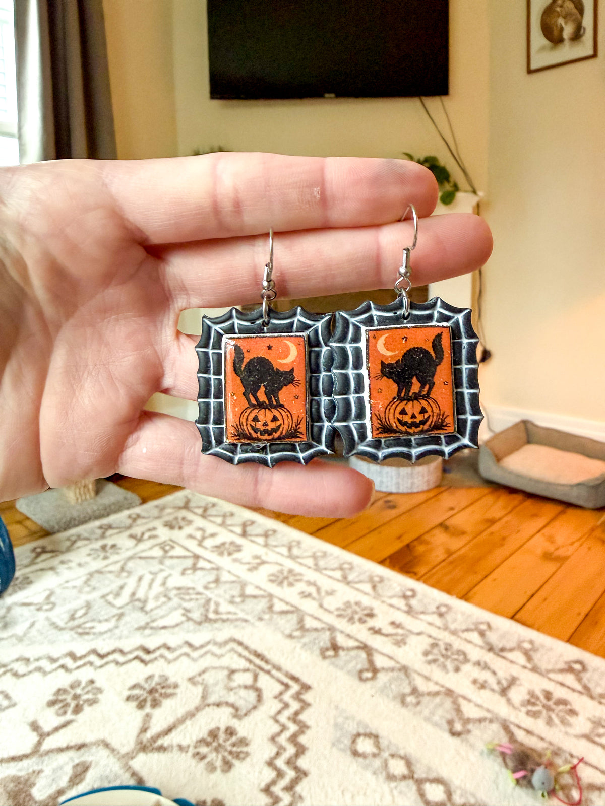 A hand holds a pair of Halloween-themed dangling earrings with orange and black cat-and-pumpkin designs, shown against a blurred living room background.