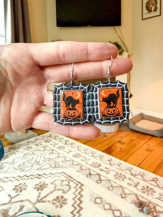 A hand holds a pair of Halloween-themed dangling earrings with orange and black cat-and-pumpkin designs, shown against a blurred living room background.