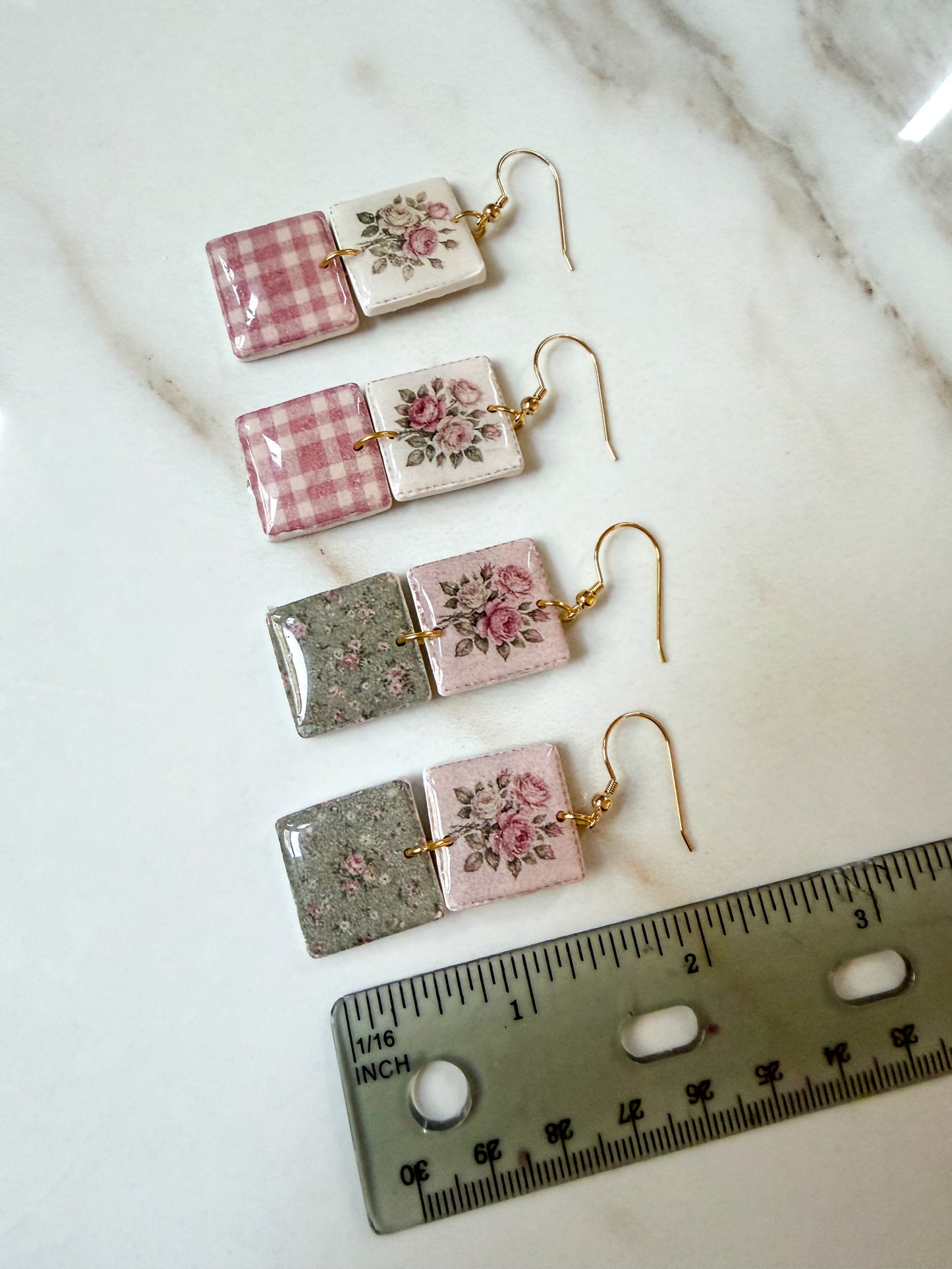 Four rectangular dangle earrings laid out on a light marble surface, with a ruler at the bottom for scale. The earrings feature mixed designs: pink gingham, pink floral, and glittery gray with floral details, all with gold-tone hooks. 2 inches in length. 