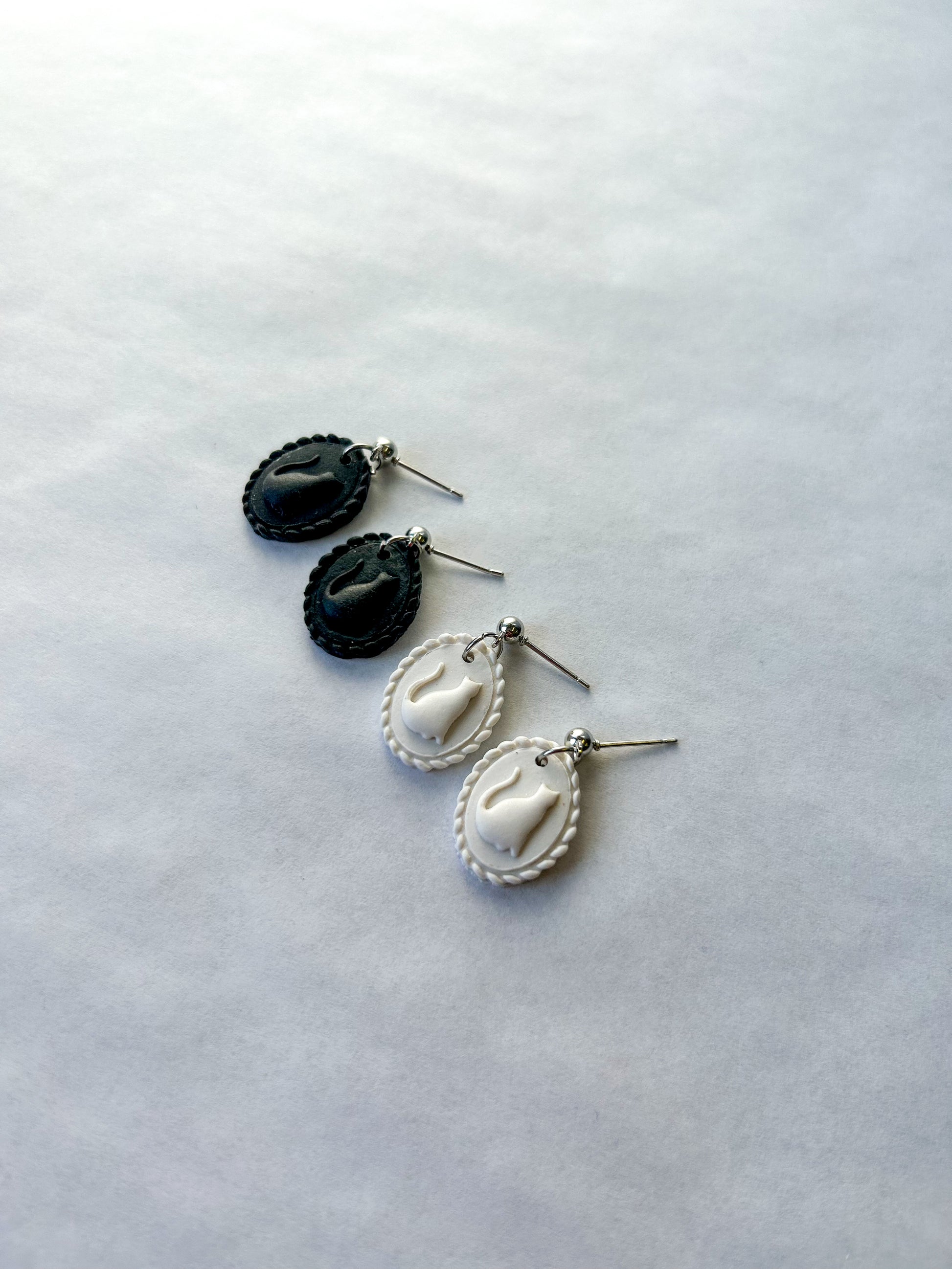 Pair of black and white earrings on a light gray background. The earrings resemble a scalloped frame with a raised cat silhouette in the center.