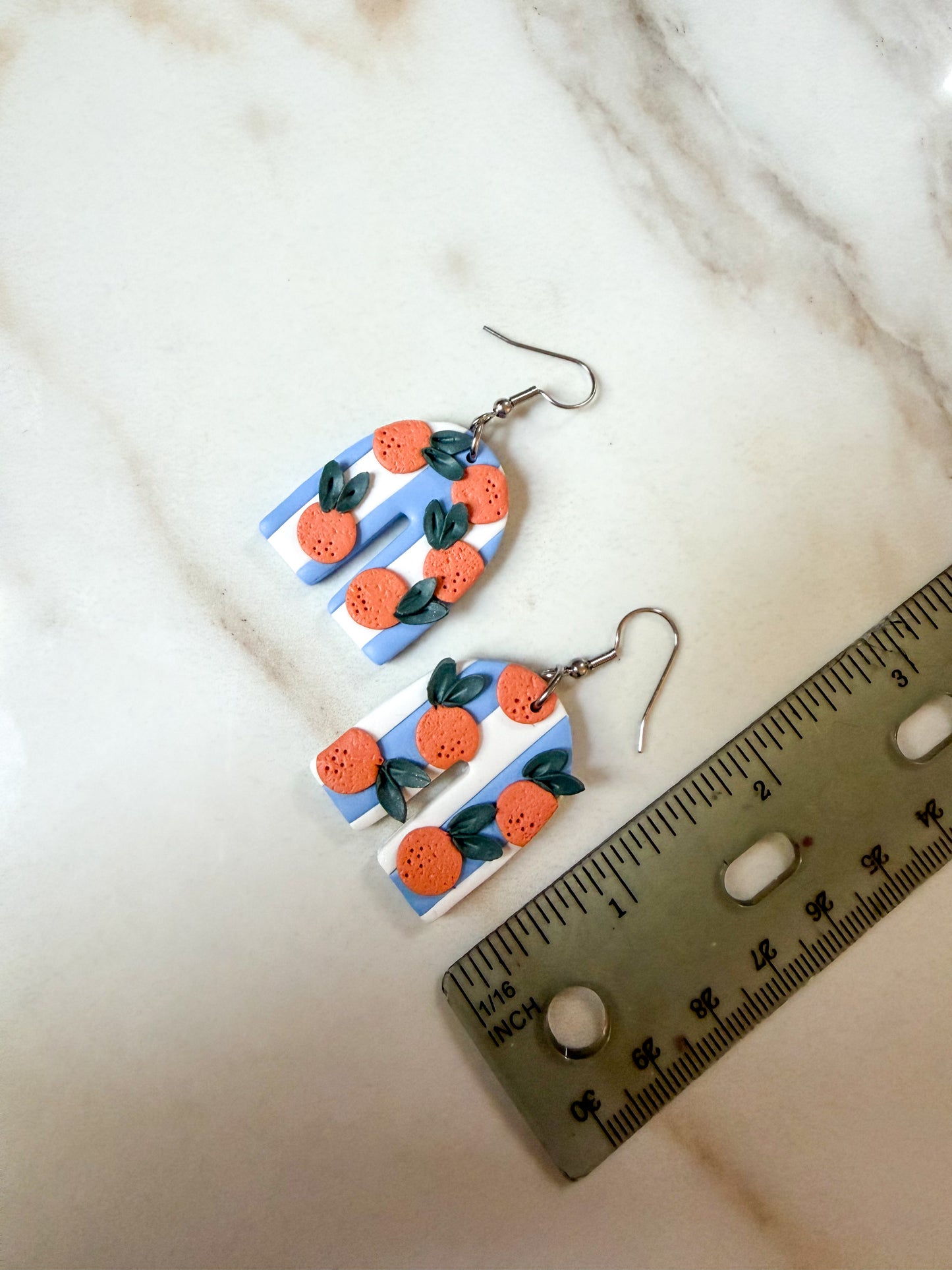 A pair of handmade polymer clay dangle earrings featuring a blue and white striped background with orange citrus fruits and green leaves, displayed on a marble surface next to a metal ruler for scale. About 2 inches long.