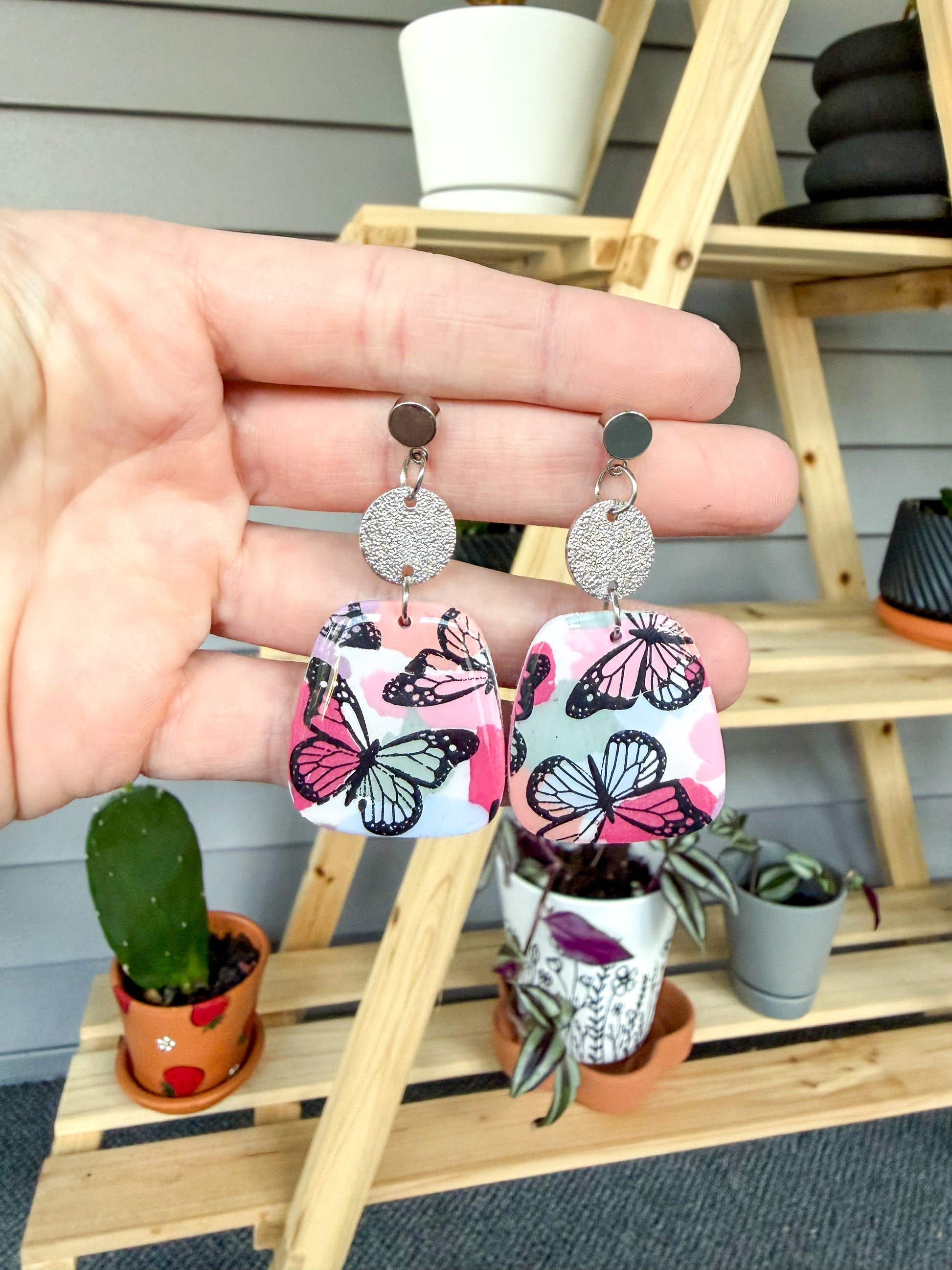 Butterfly-patterned earrings held by a hand with a wooden shelf and plants in the background.