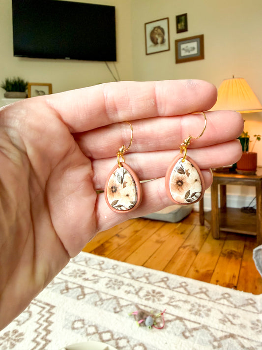 A hand holds a pair of teardrop-shaped earrings with a floral pattern on a cream background, against a blurred living room background.