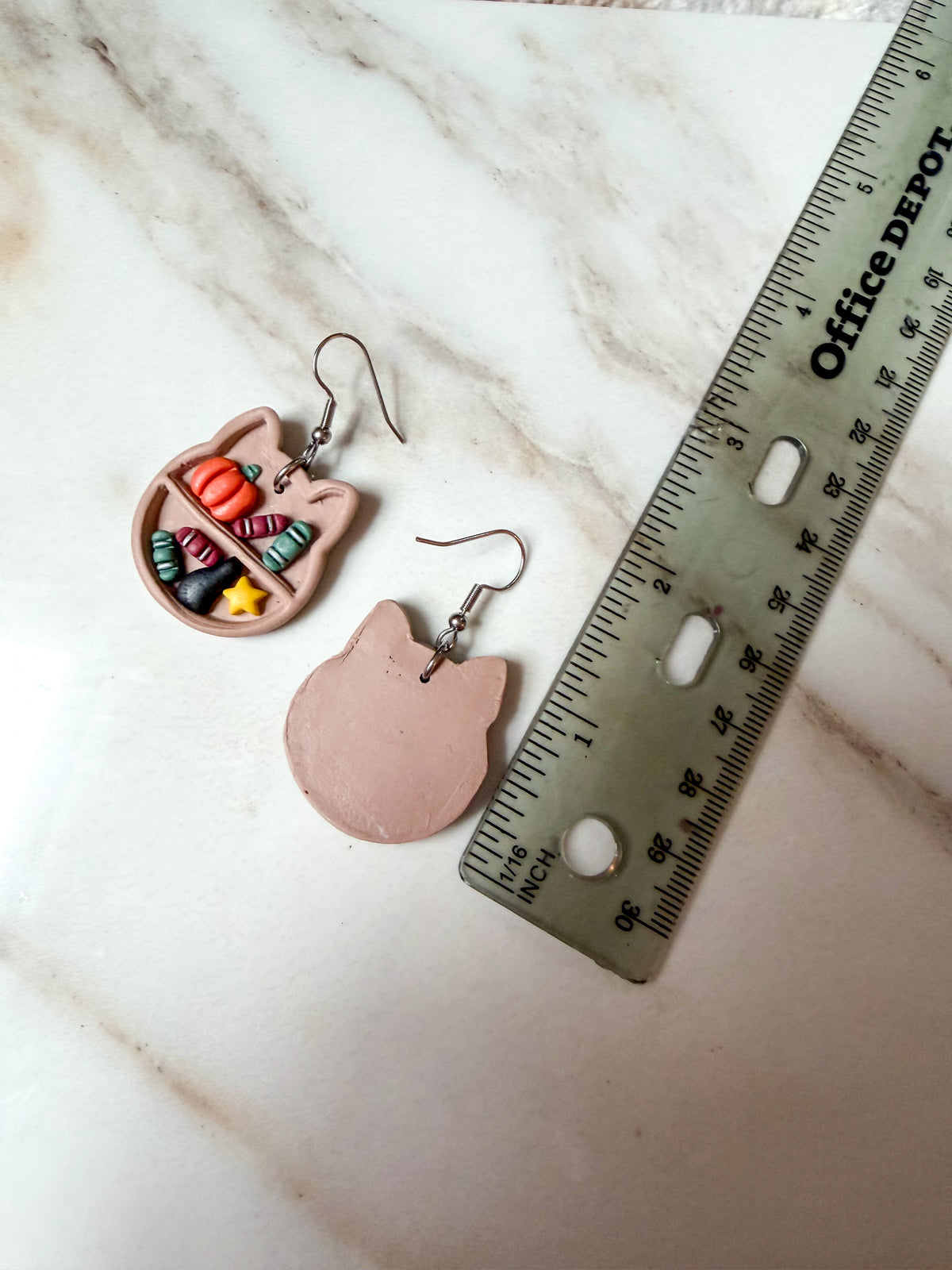 Cat head bookshelf earrings laying on a flat marble surface with a ruler for scale. About 2 inches in length. 
