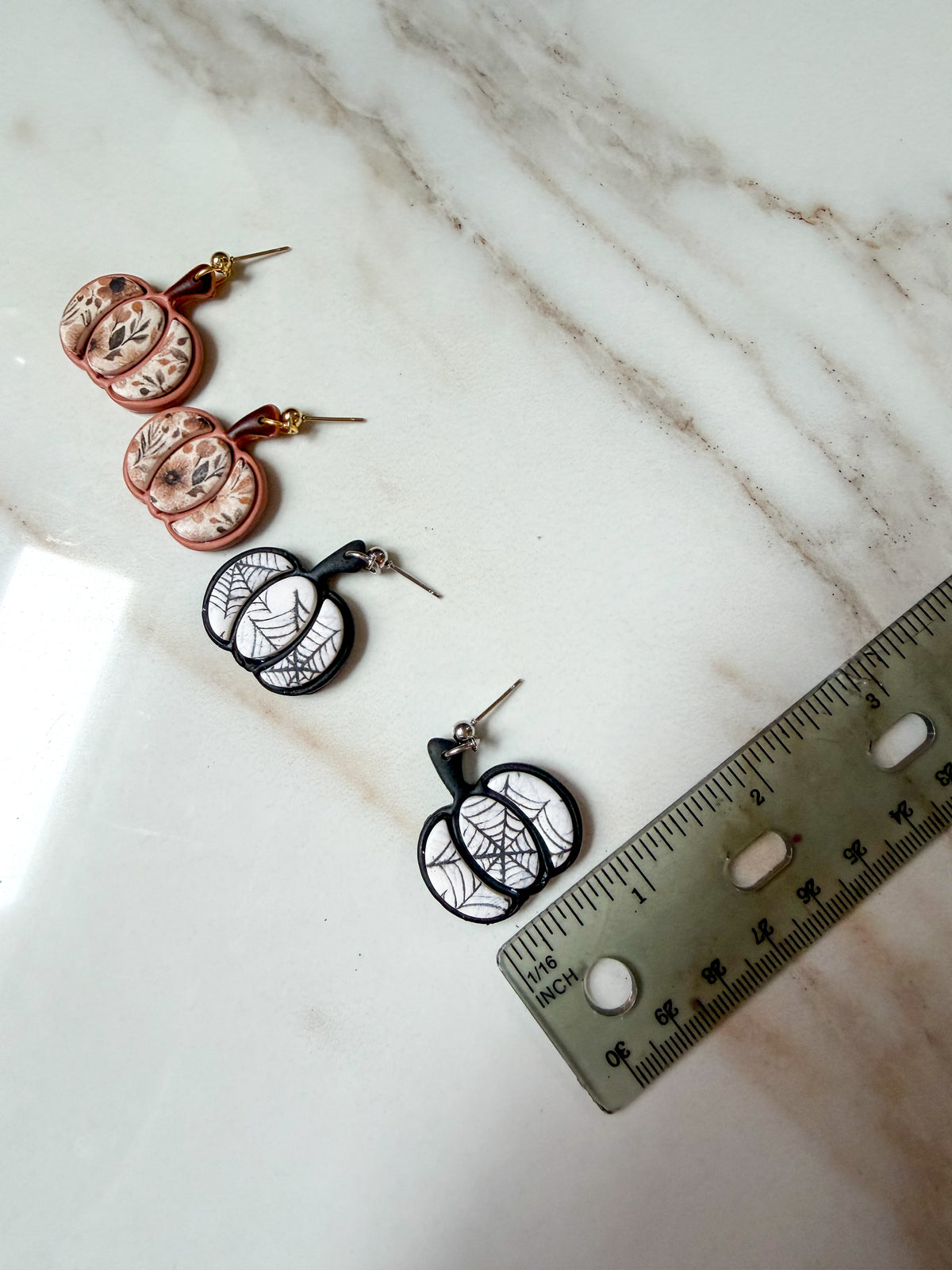 Two pairs of Halloween-themed earrings displayed on a white marble surface next to a ruler for scale. One pair features round pinkish-beige florals and another features spiderweb patterns. Around one inch in width.