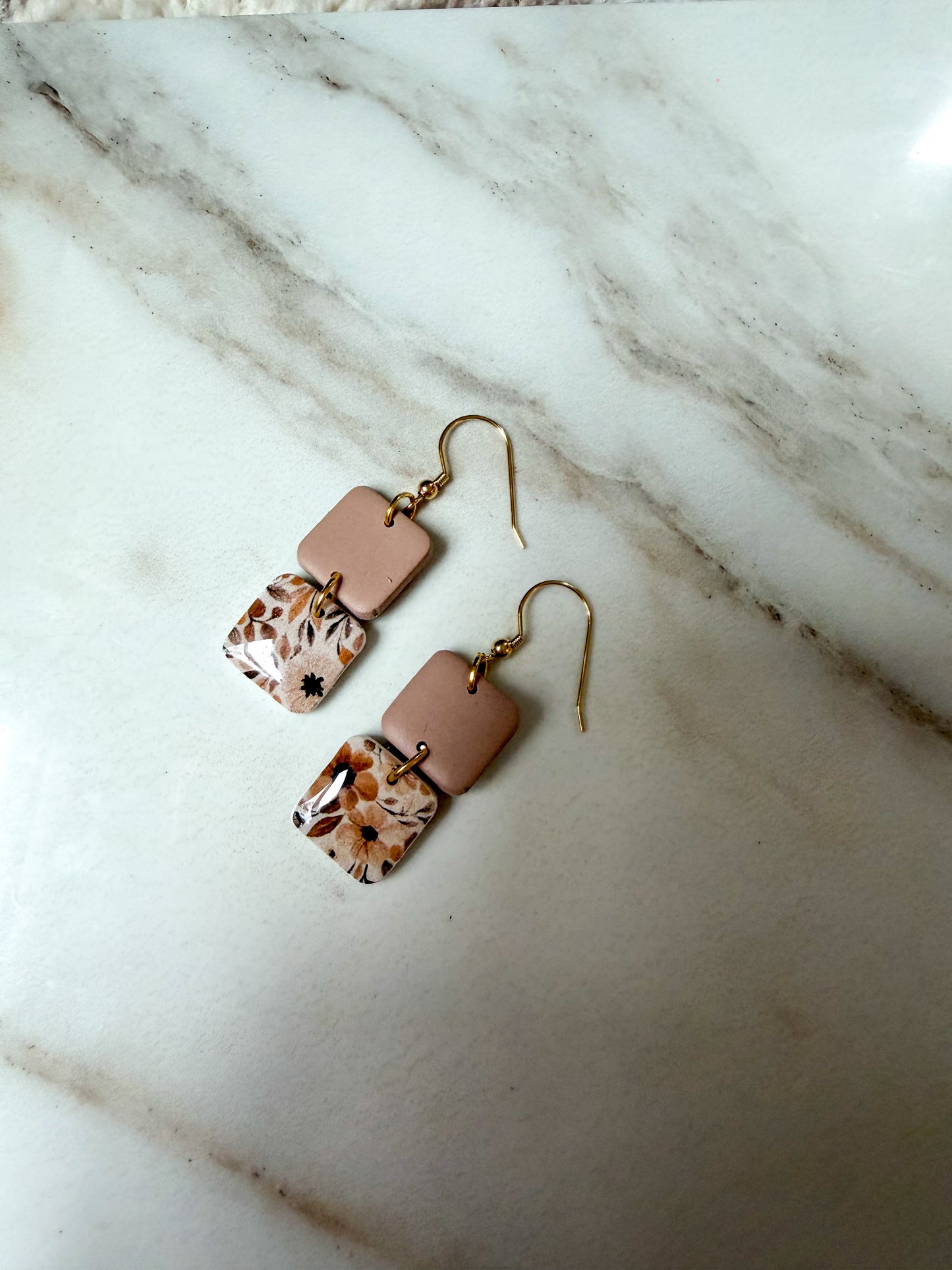 A pair of handmade dangle earrings with brown rectangular pieces, patterned square drops in orange, brown, and cream, and gold-tone fishhook wires, displayed on a white marble surface.