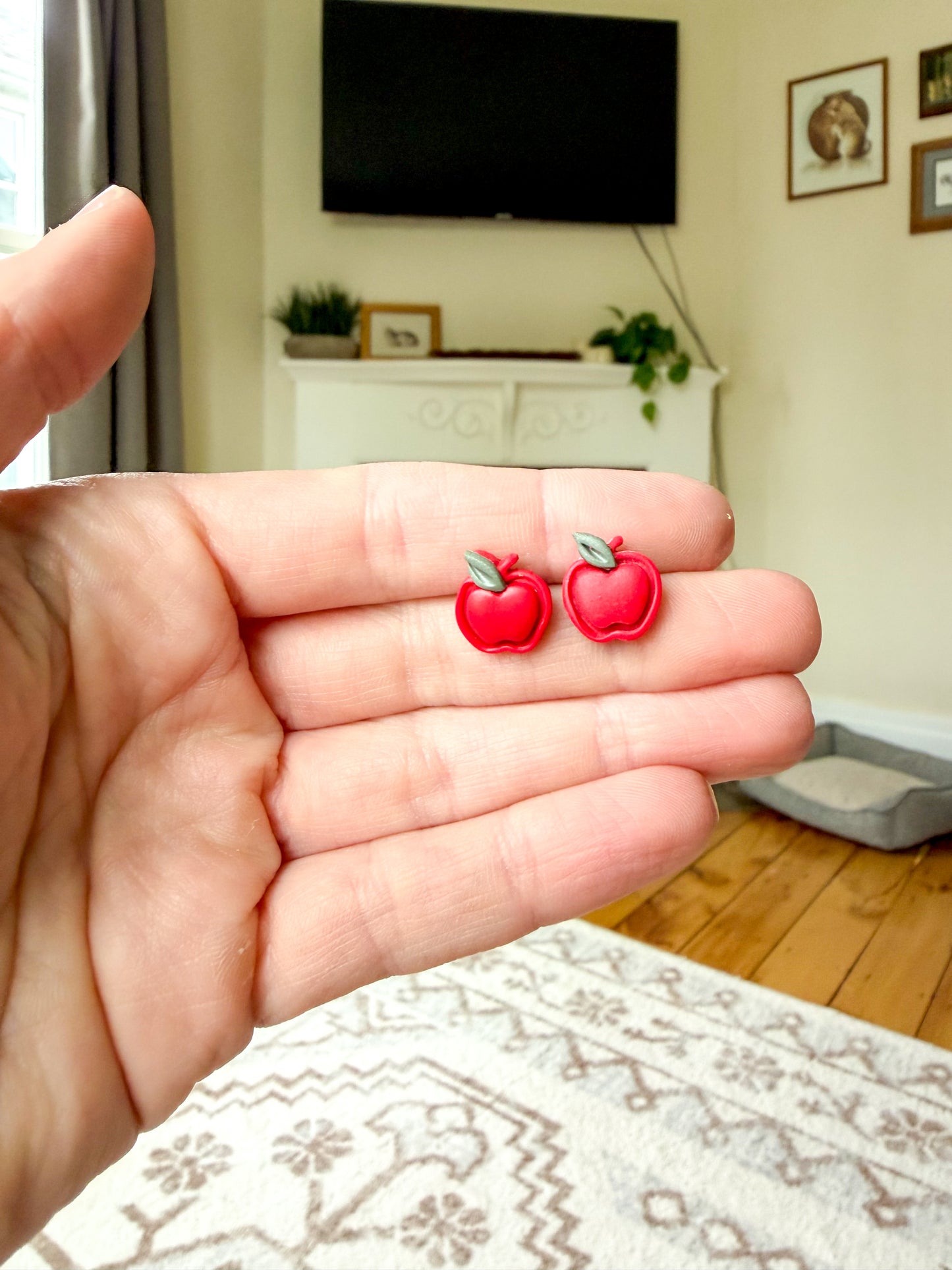 A hand holding a pair of apple-shaped earrings against a living room background. 
