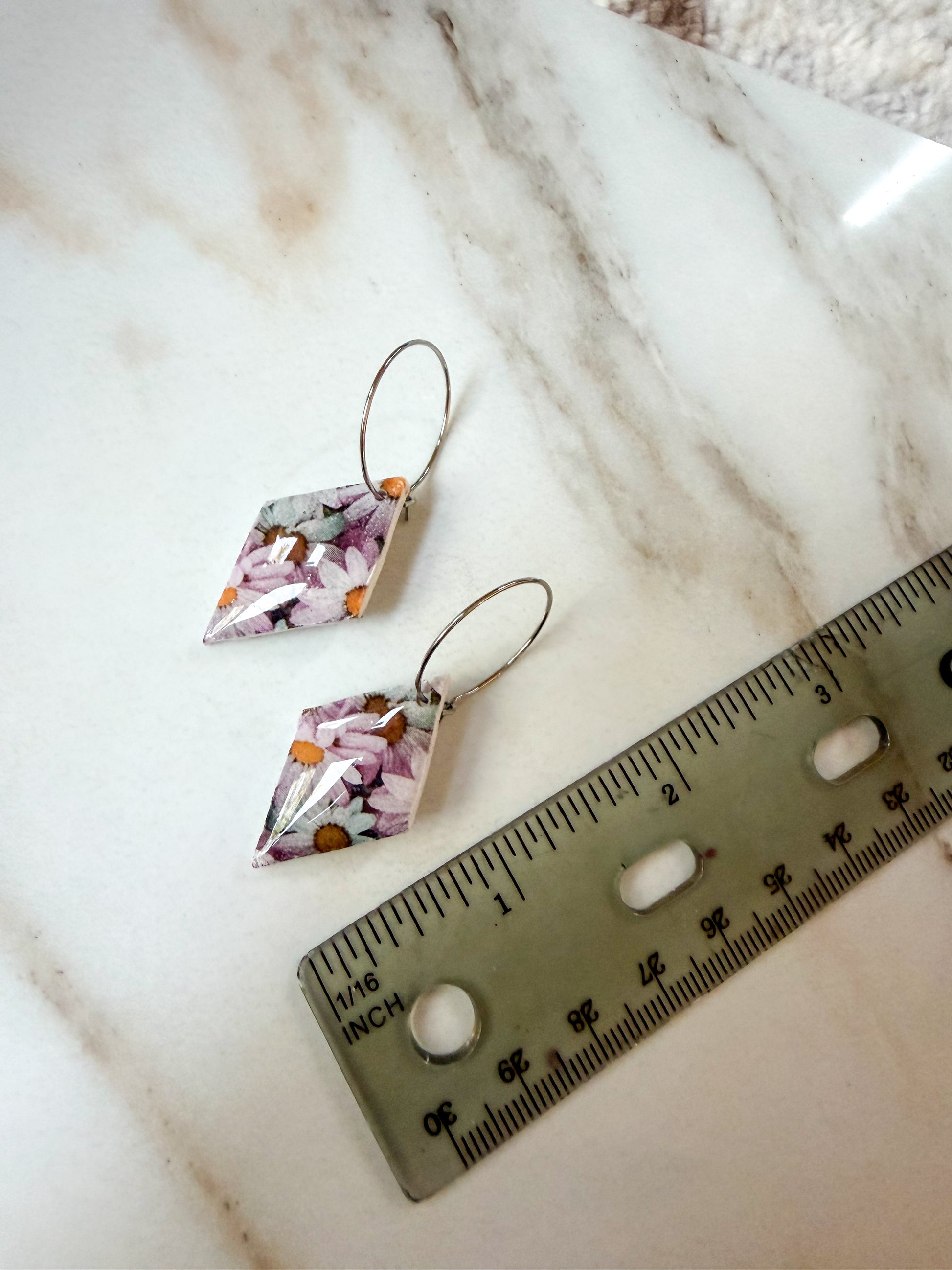 A pair of dangling earrings with irregular diamond-shaped floral resin pendants and silver-tone wire hoops, shown on a white marble surface next to a ruler for scale. About 2 inches in length. 
