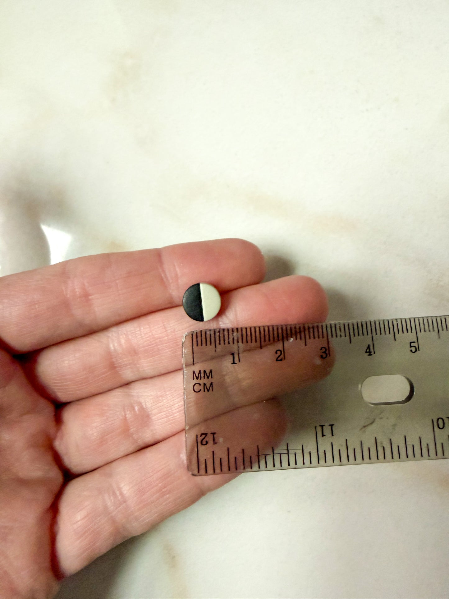 Small circle stud held between fingers in front of a ruler for scale. About 3/4 cm in length 