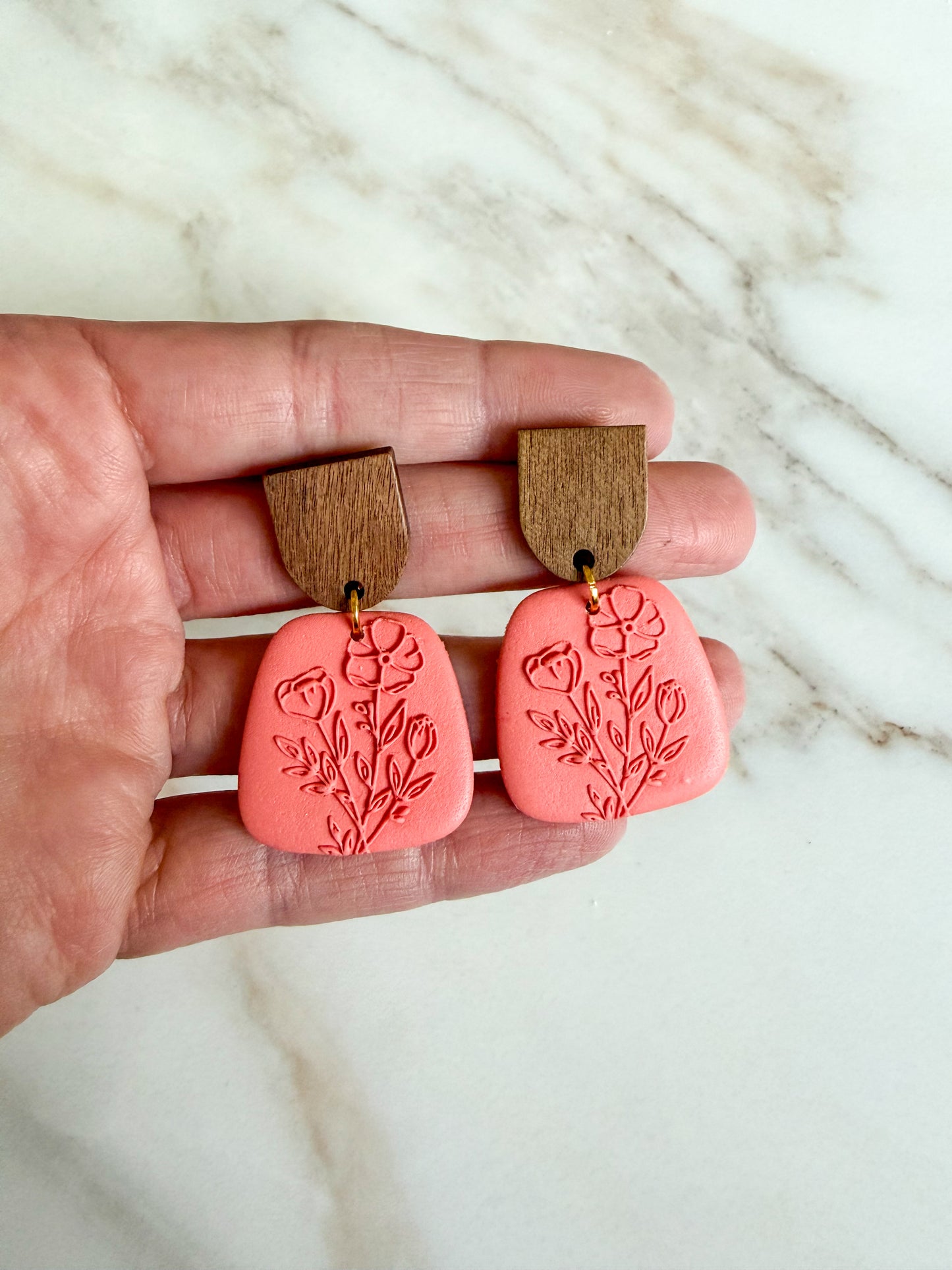 Pink earrings with wooden tops held between fingers on a marble background