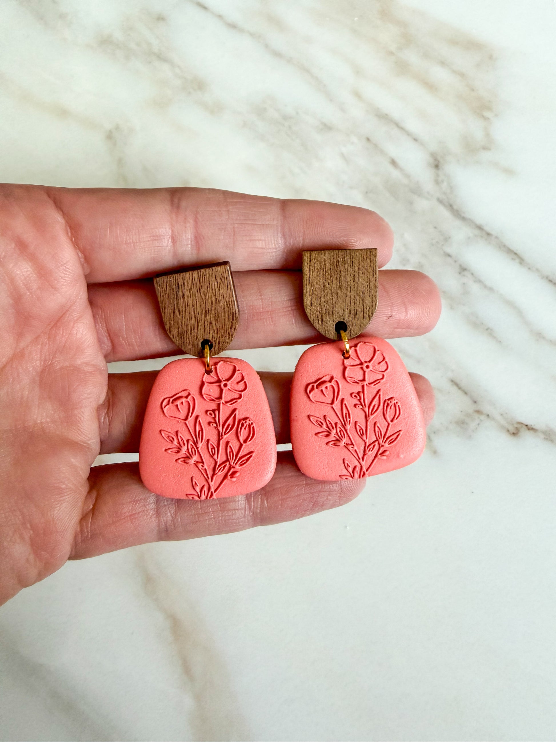 Pink earrings with wooden tops held between fingers on a marble background