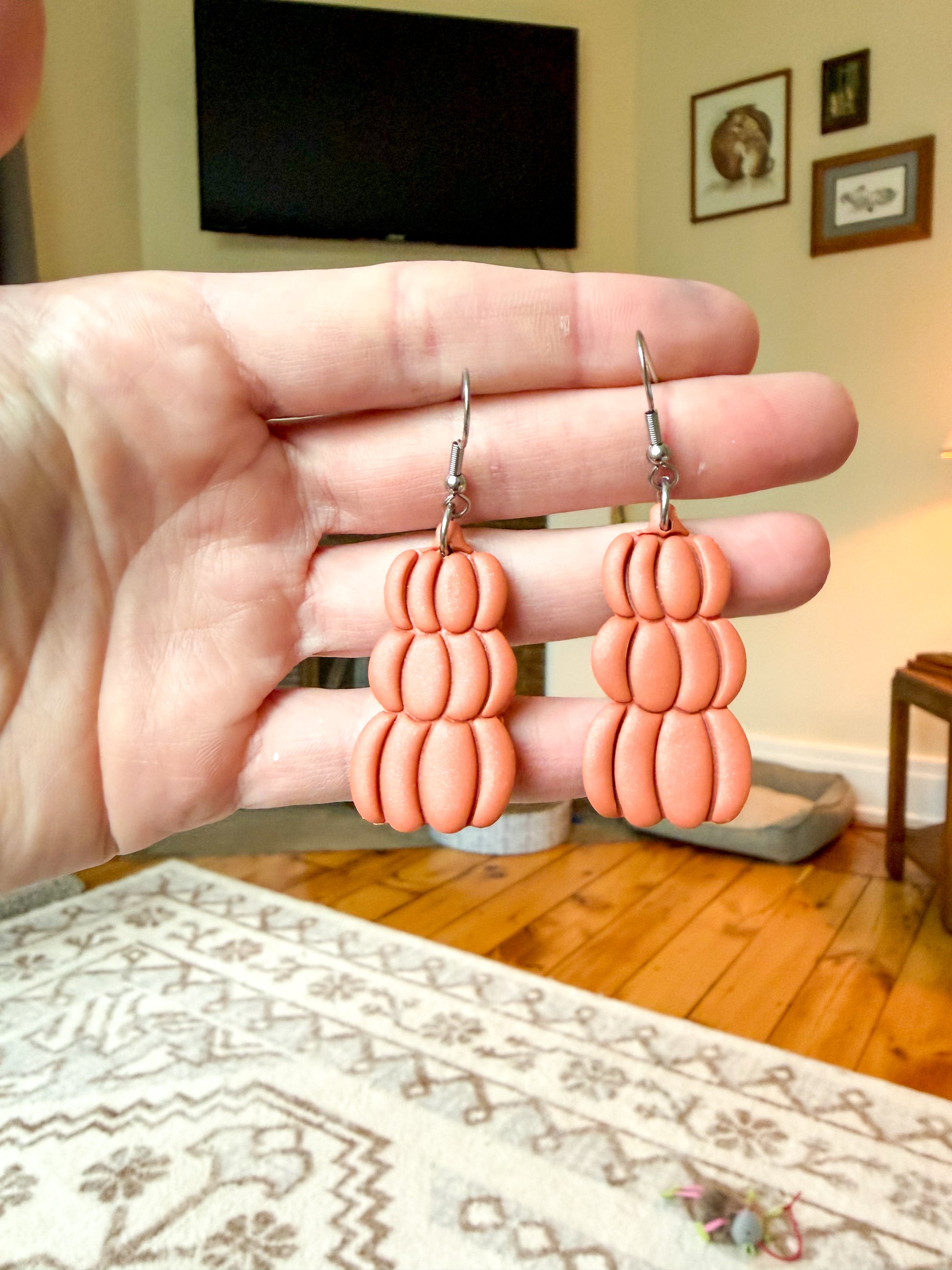 A pair of pumpkin dangle earrings held by a person's hand in a home setting with a wooden floor and framed pictures in the background.