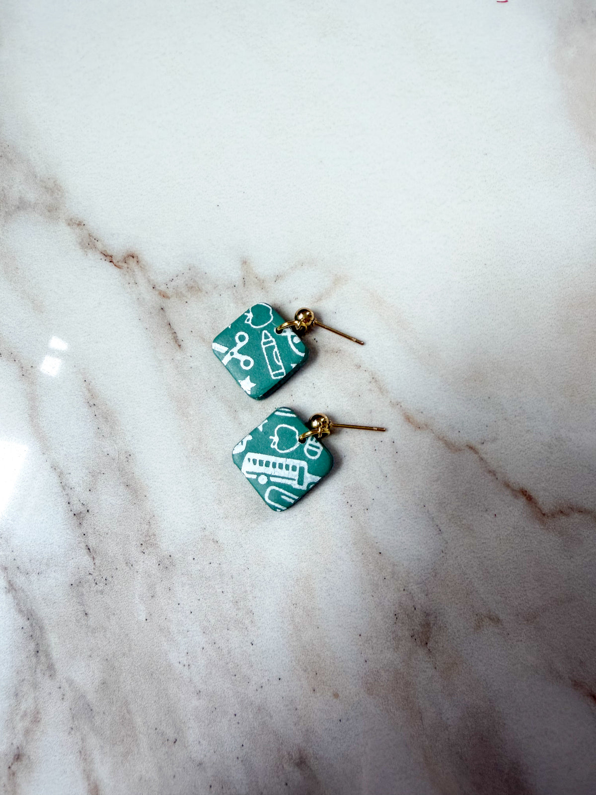 A pair of square green and white patterned dangle earrings with gold-tone posts, resting on a white marble surface.