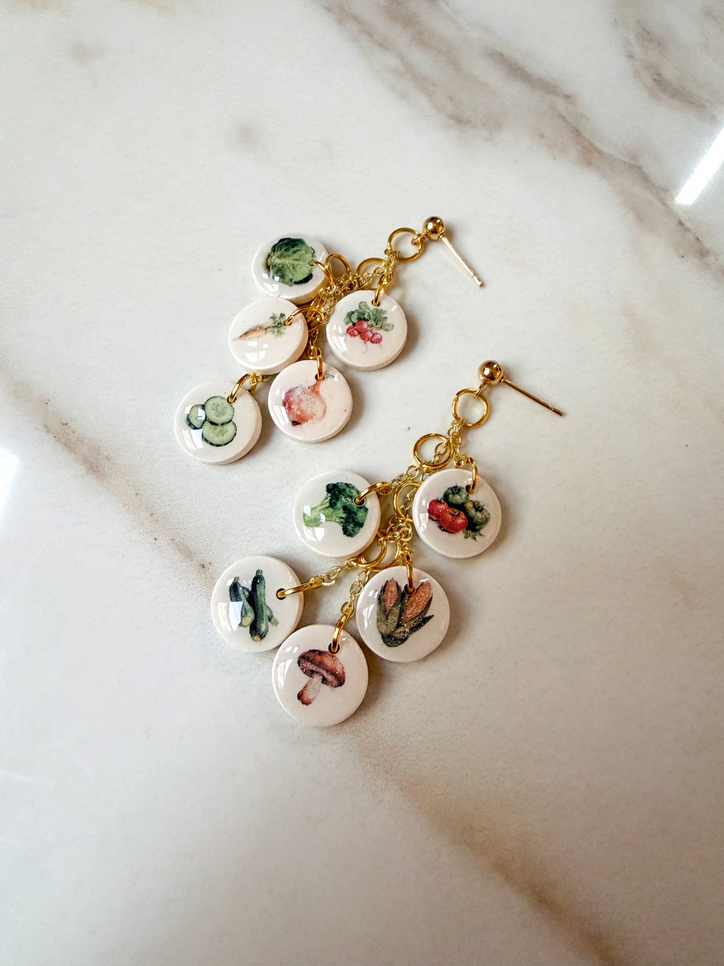 A pair of gold-tone dangle earrings with multiple small round white charms printed with colorful vegetable illustrations, displayed on a light marble surface.