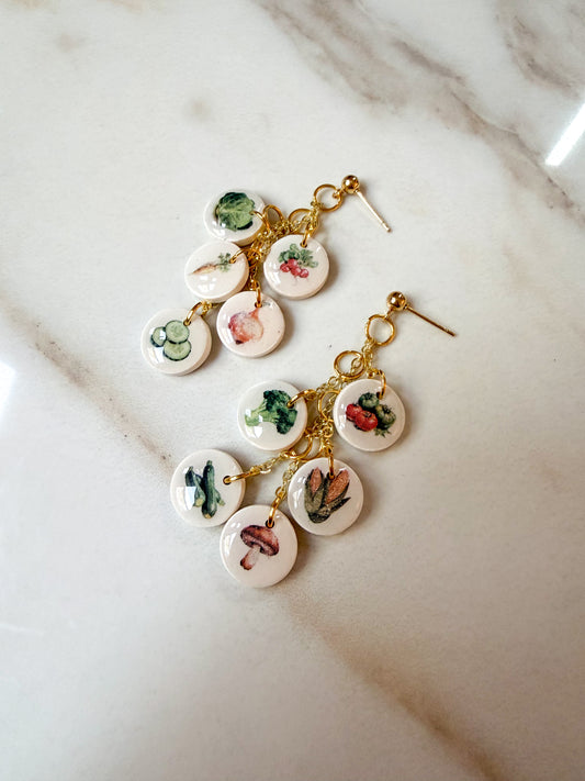 A pair of gold-tone dangle earrings with multiple small round white charms printed with colorful vegetable illustrations, displayed on a light marble surface.