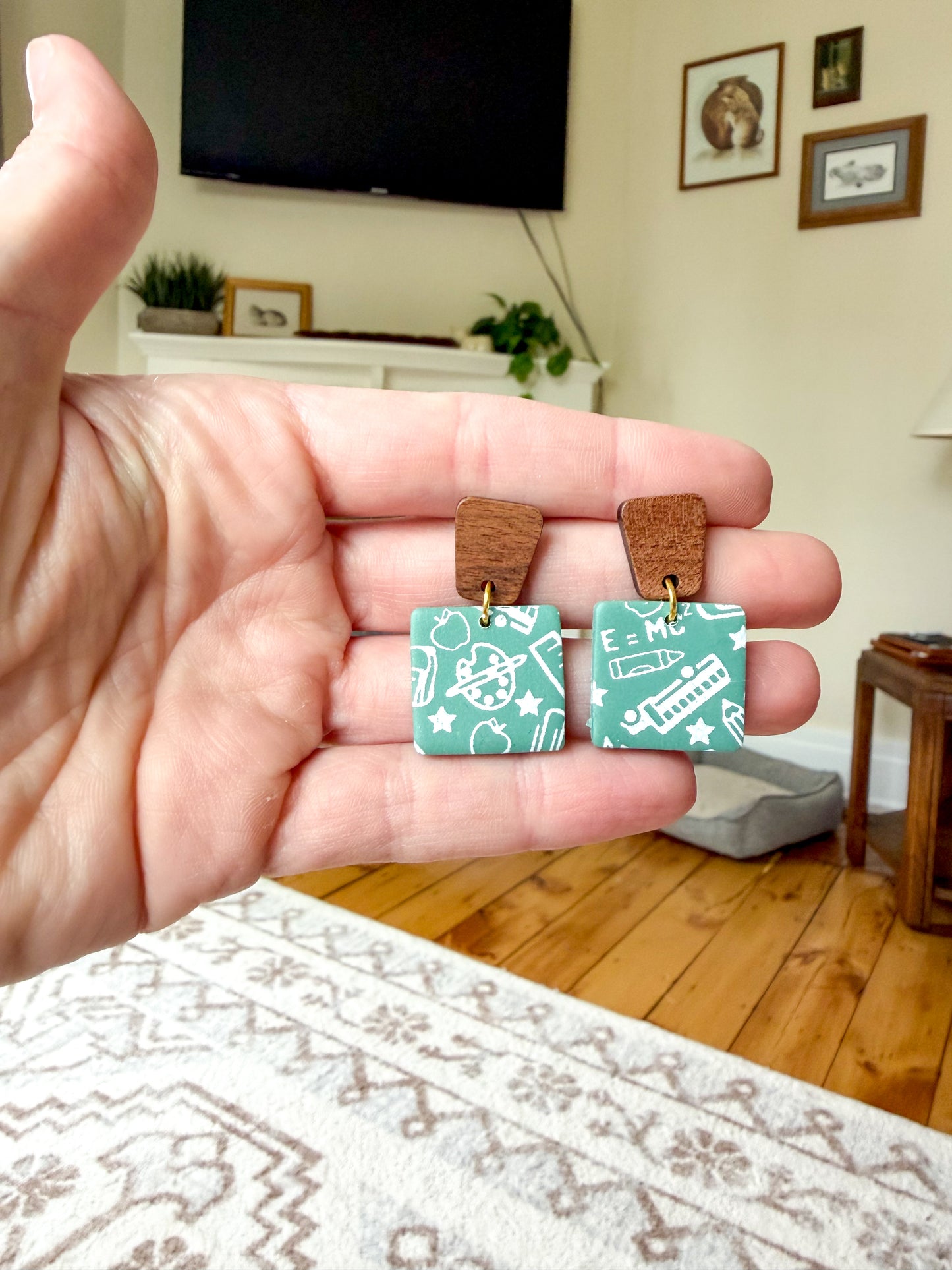 A hand holding a pair of earrings against a living room background. They are green to resemble old chalkboards, with white school-themed scribble designs. The posts are wood. 