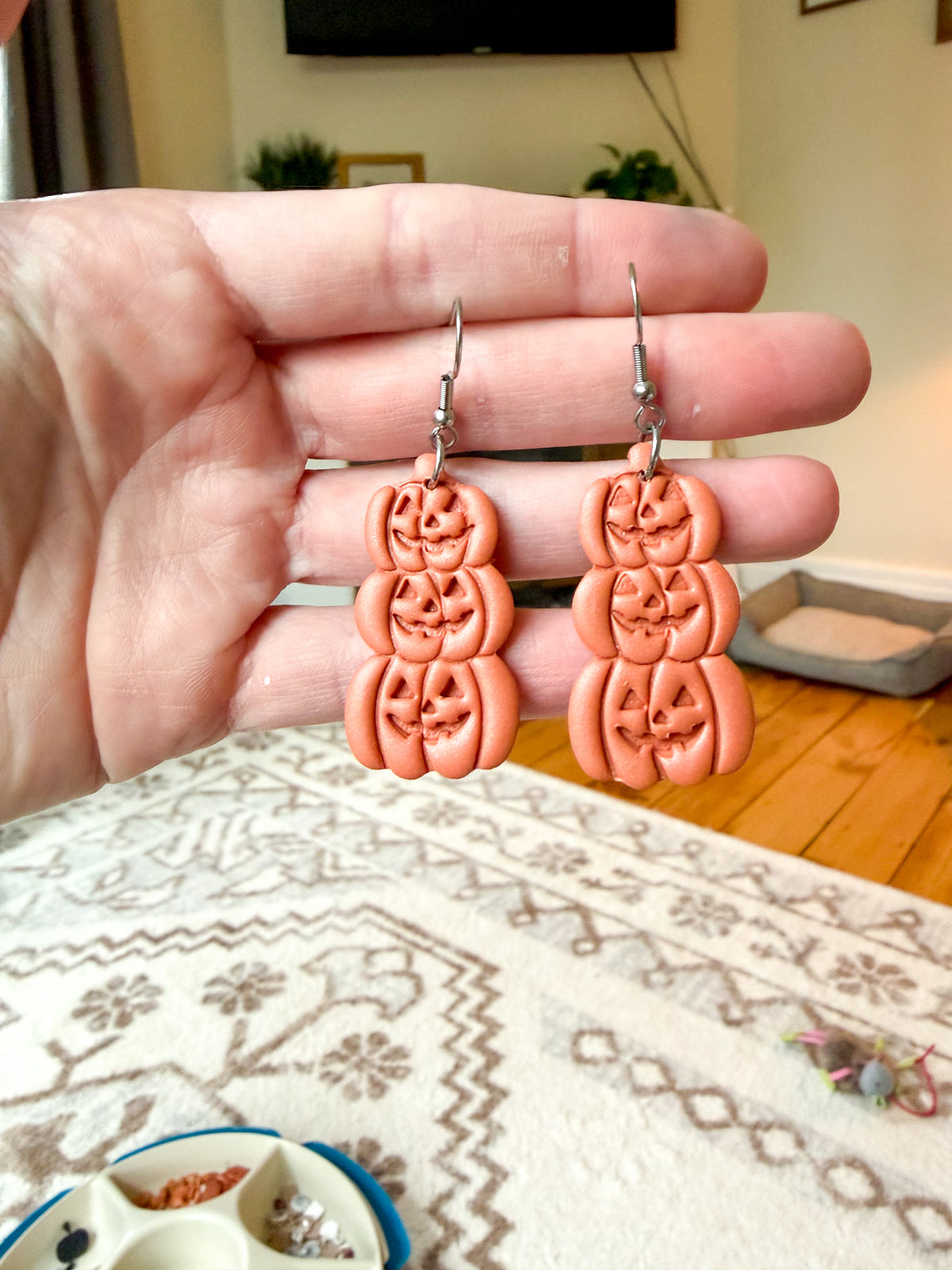A pair of handmade jack-o-lantern drop earrings held up by a person's hand, with a home interior background visible.