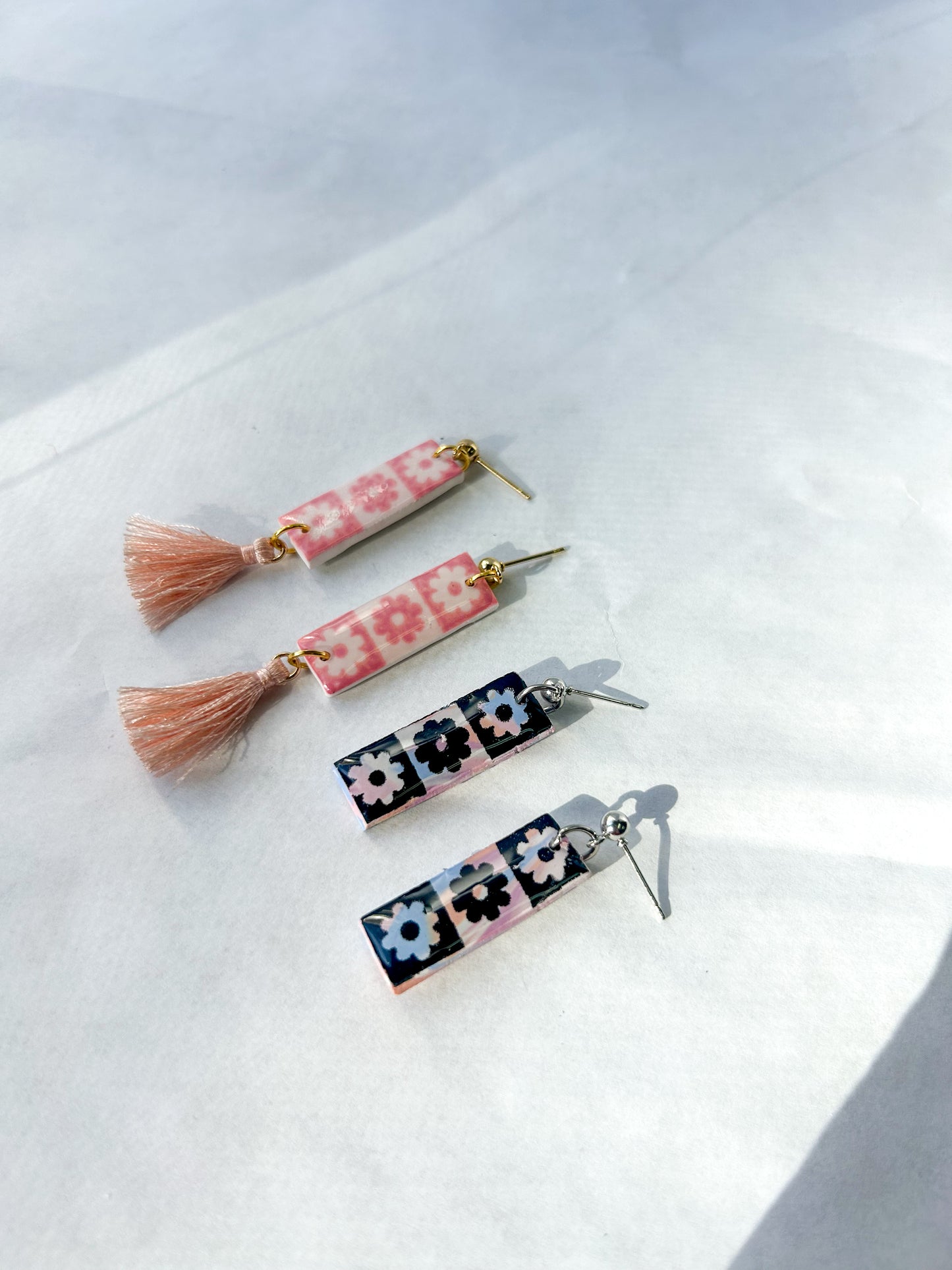 Two pairs of earrings flying on a flat surface. One pair is pink checkered with a pink tassel. One pair is black checker with no tassel