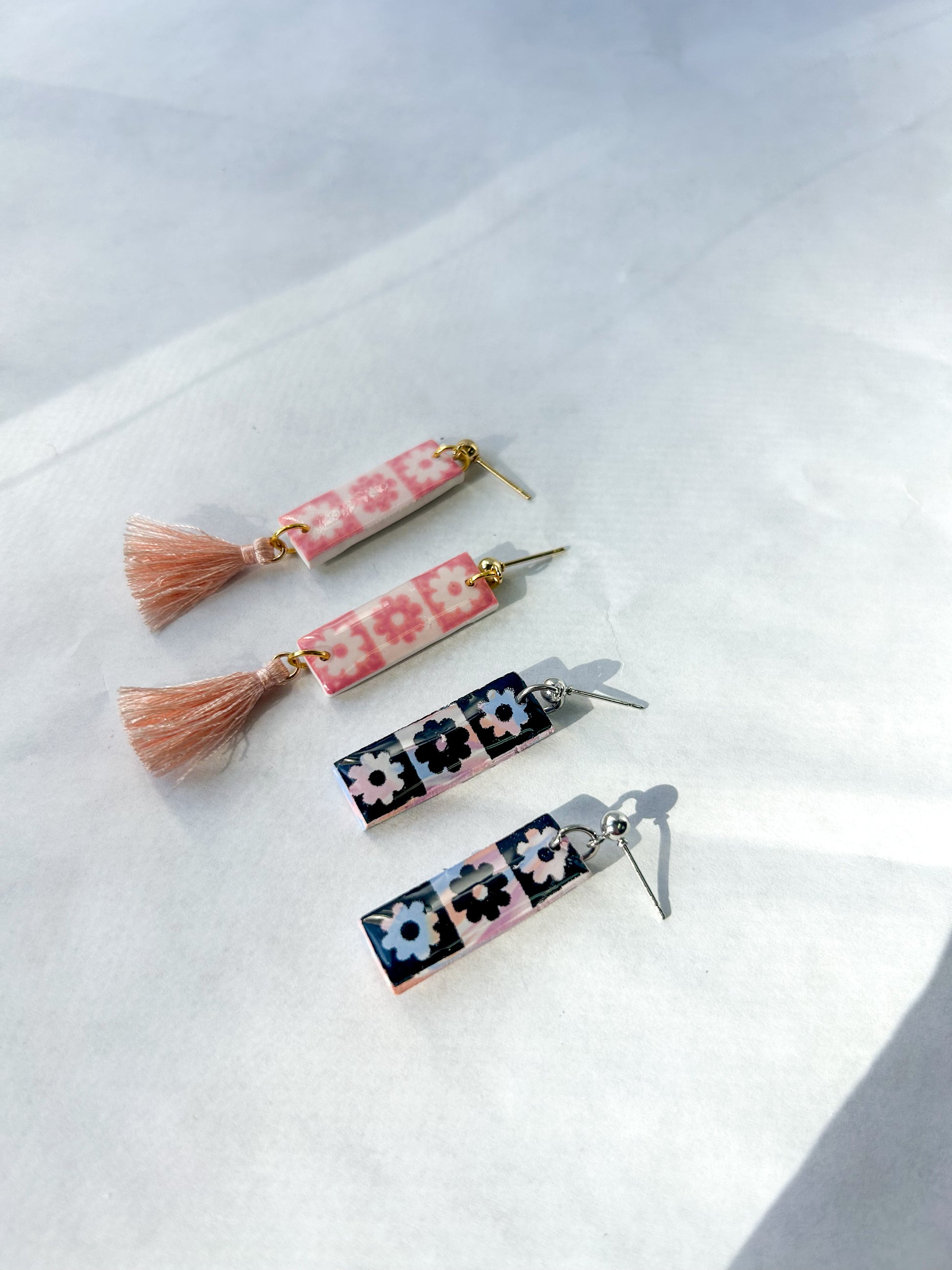 Two pairs of earrings flying on a flat surface. One pair is pink checkered with a pink tassel. One pair is black checker with no tassel