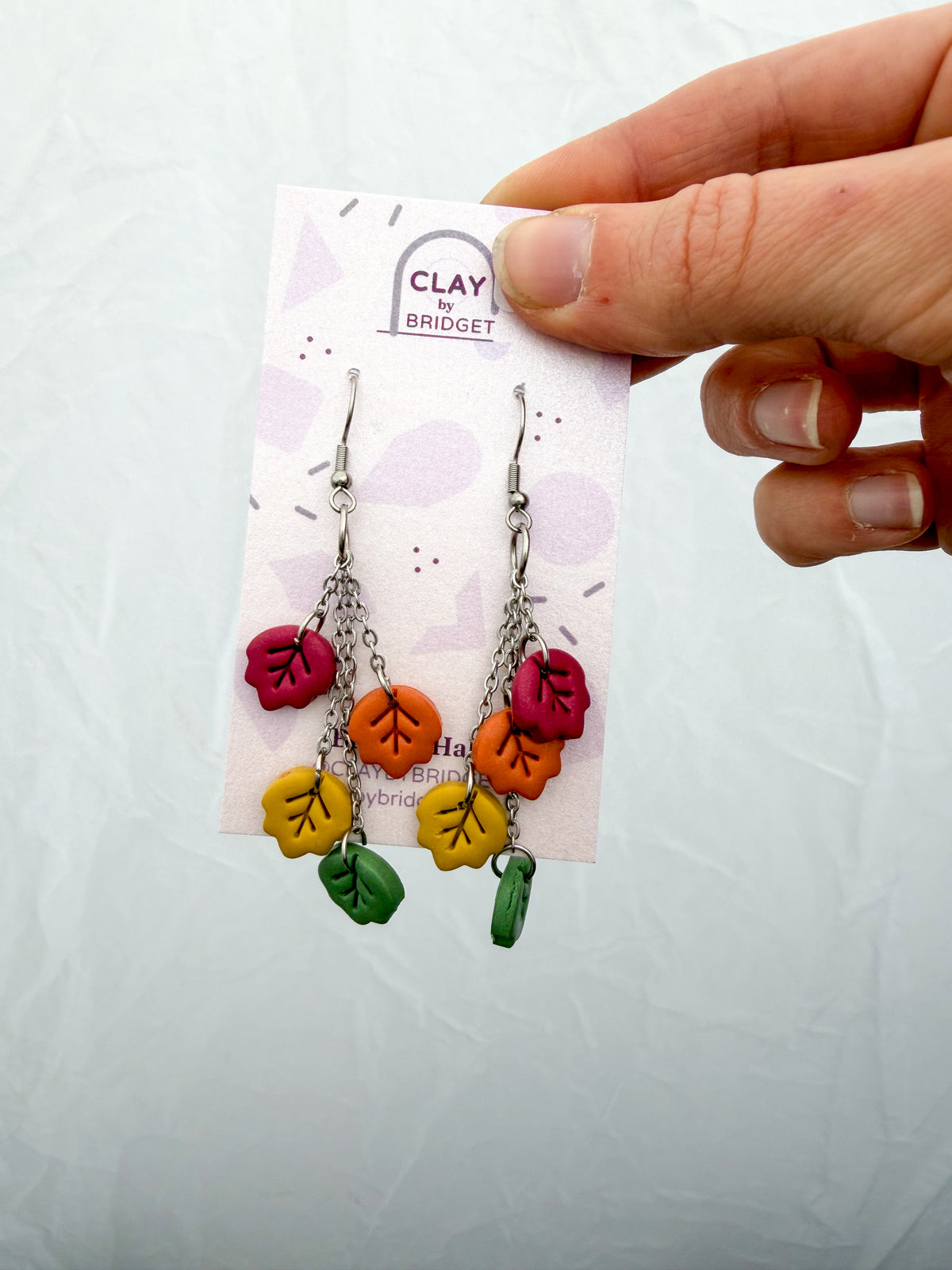 A pair of handmade clay dangle earrings with autumn leaf charms in red, orange, yellow, and green, displayed on a branded backing card held by a hand.