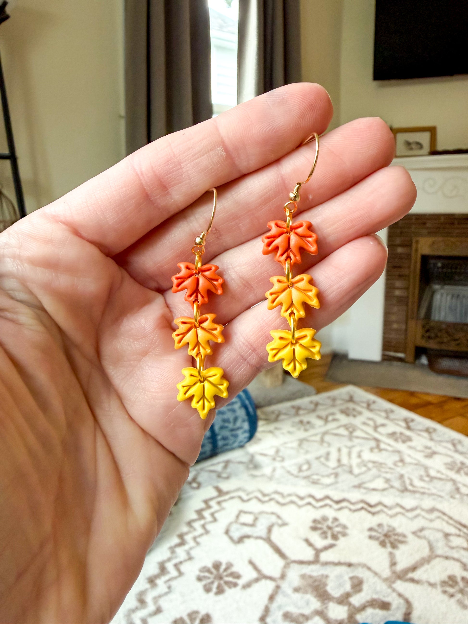 A hand holds a pair of dangling earrings shaped like autumn leaves in orange, yellow, and orange-yellow tones with gold hooks, against a blurred indoor background.