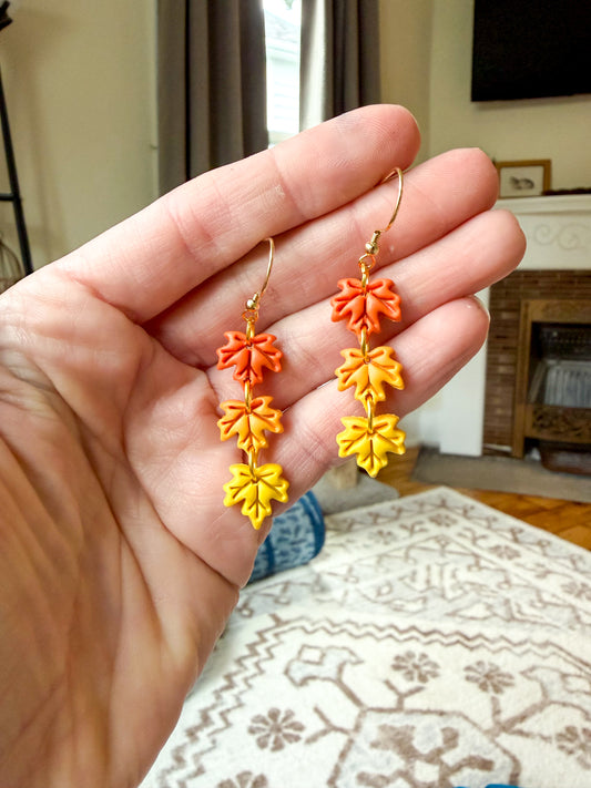 A hand holds a pair of dangling earrings shaped like autumn leaves in orange, yellow, and orange-yellow tones with gold hooks, against a blurred indoor background.