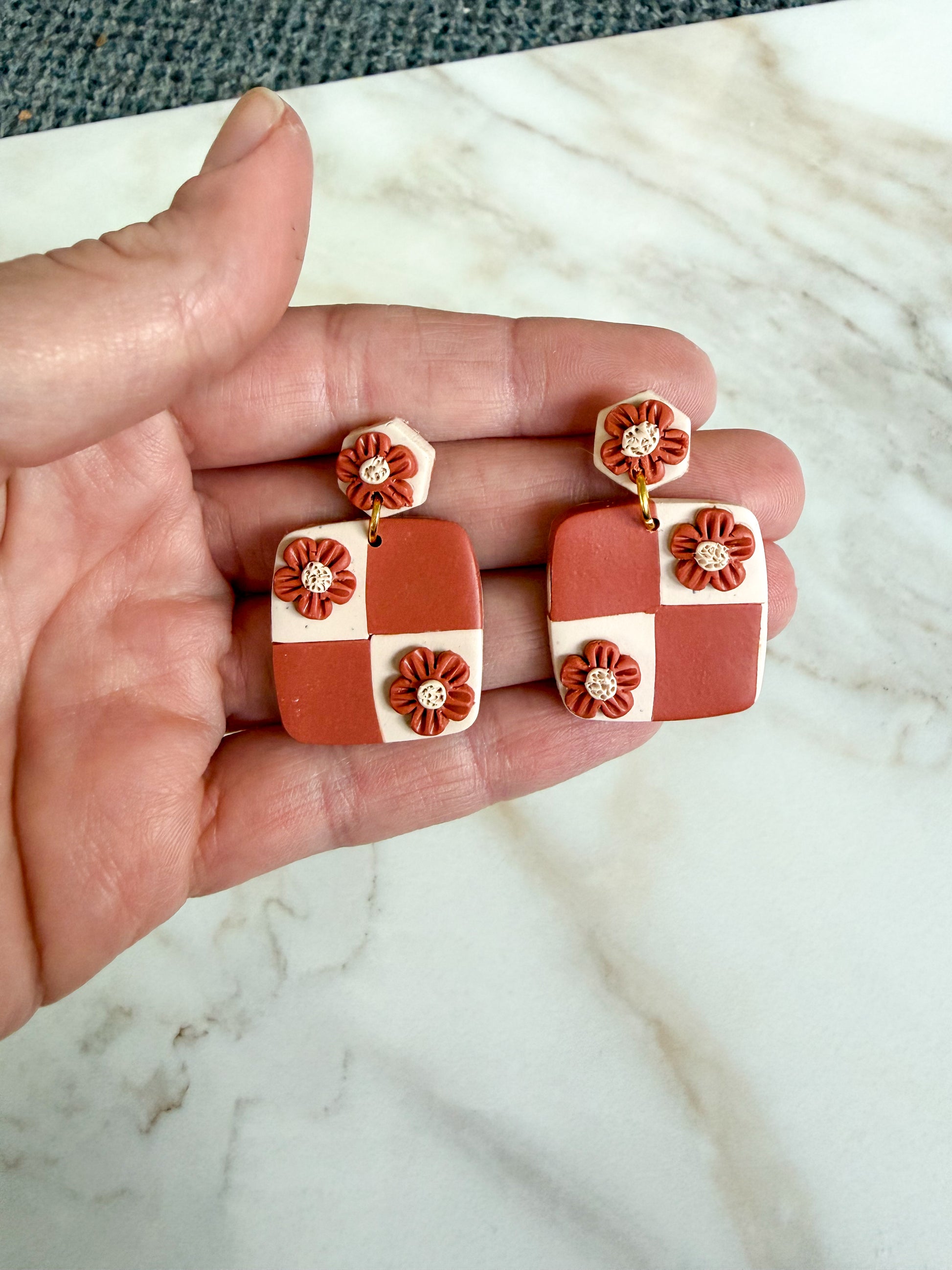 Hand holding a pair of terracotta and white checkered earrings on a marble surface