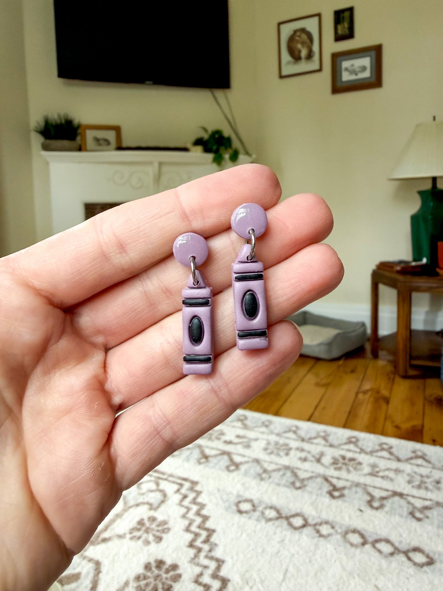 A hand holds a pair of purple polymer clay earrings shaped like crayons, with black and silver details.