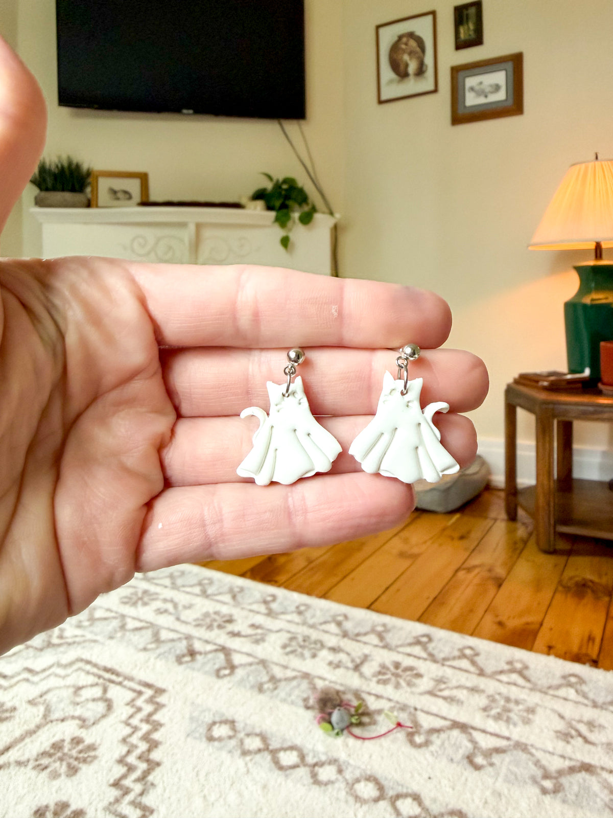 A hand holds a pair of white cat-shaped dangle earrings with silver-toned hooks against a blurred living room background.