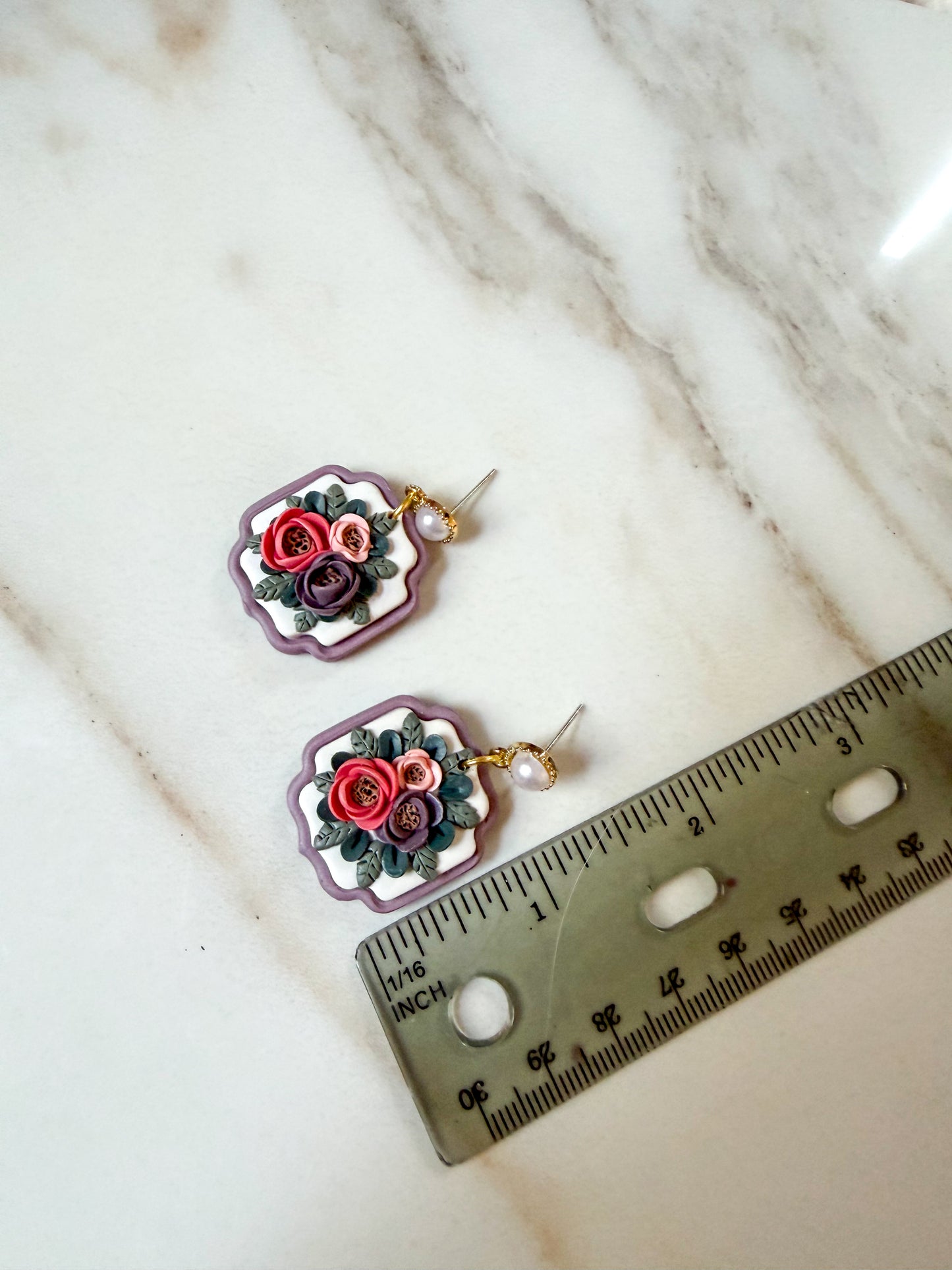 A pair of handmade floral earrings with purple and pink flowers, green leaves, and small pearl-like beads, displayed on a white marble surface next to a metal ruler for scale. They are about 1 1/2 inches long. 