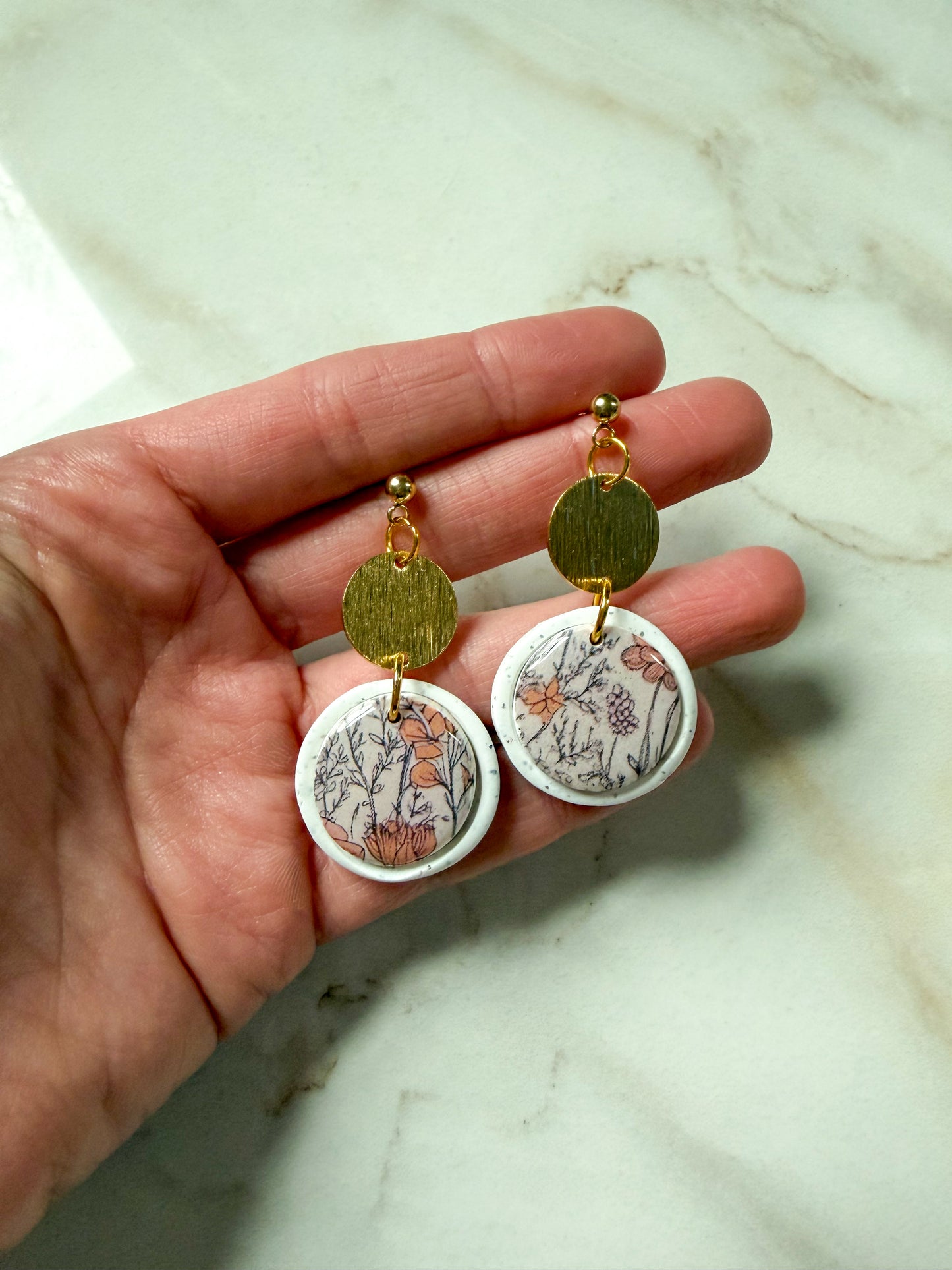 Hand holding a pair of earrings with floral designs on a marble surface
