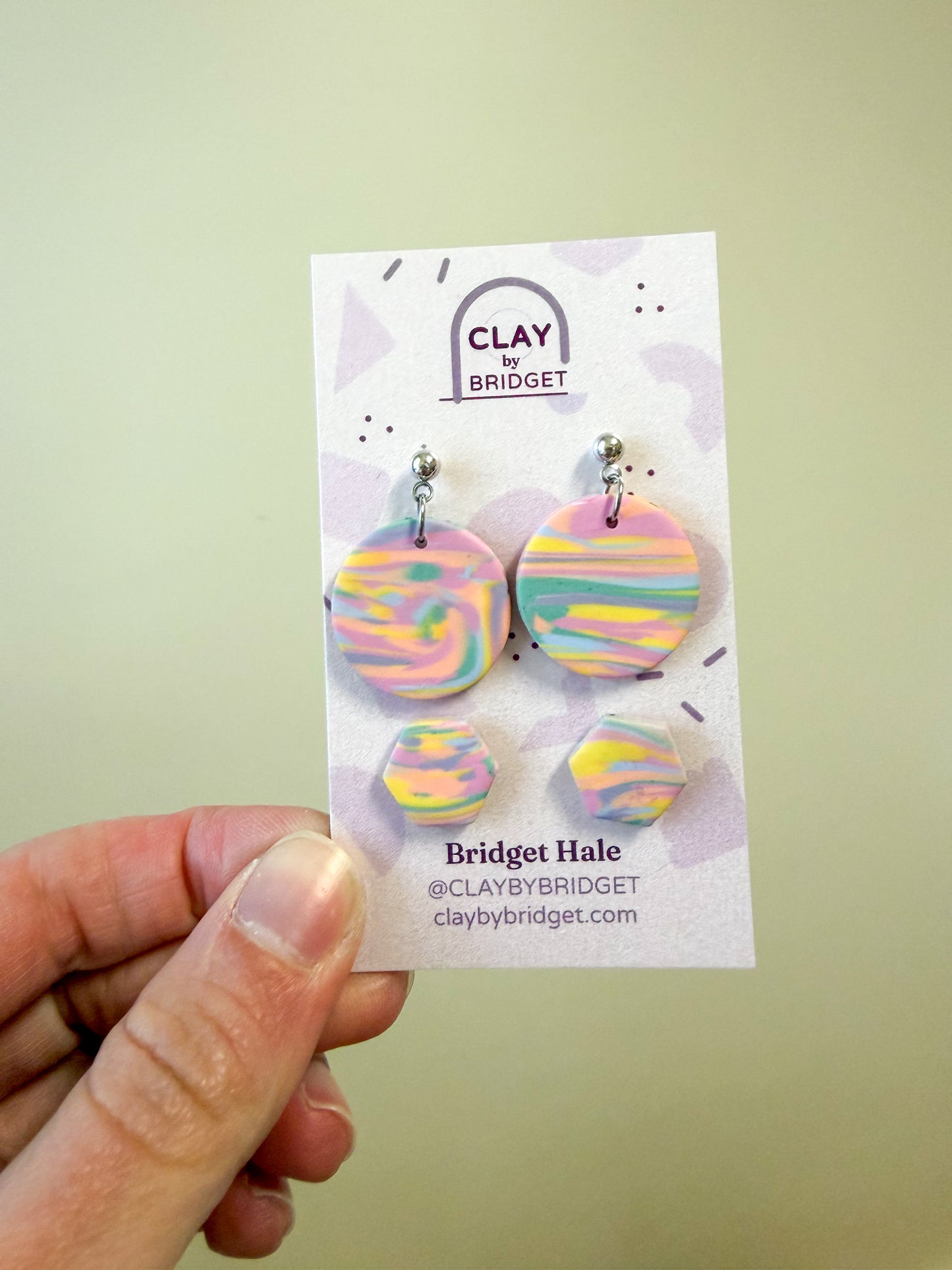 A hand holds a display card featuring four round dangle earrings with pastel marbled swirls in pink, yellow, blue, and green. The card is branded 'Clay by Bridget' and shows the name 'Bridget Hale' with social handle and website.