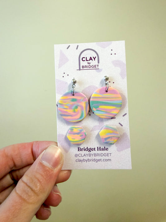 A hand holds a display card featuring four round dangle earrings with pastel marbled swirls in pink, yellow, blue, and green. The card is branded 'Clay by Bridget' and shows the name 'Bridget Hale' with social handle and website.