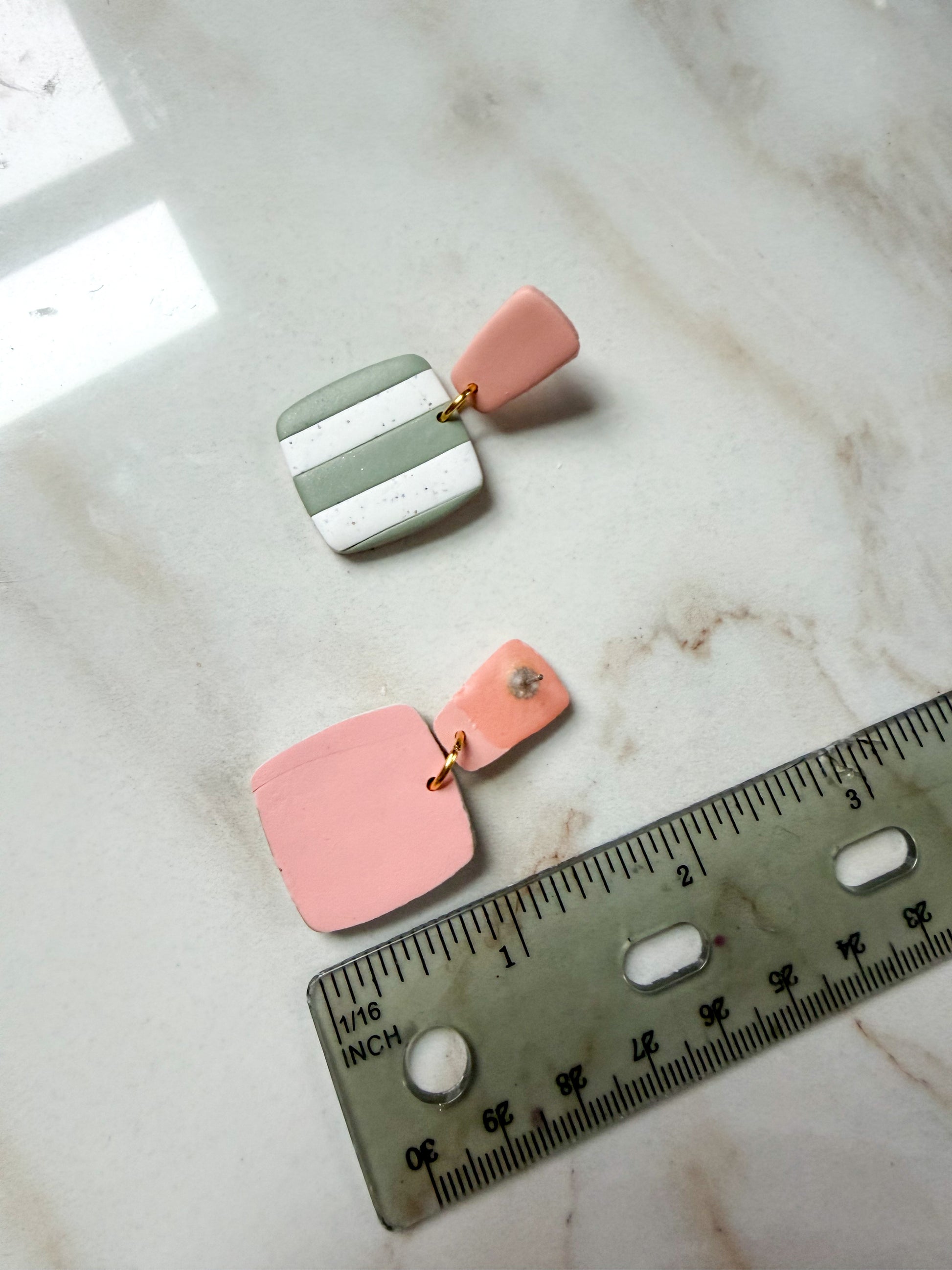 Pair of earrings with green and white striped design with pink posts on a marble surface with a ruler for scale. Just under 2 inches long. 