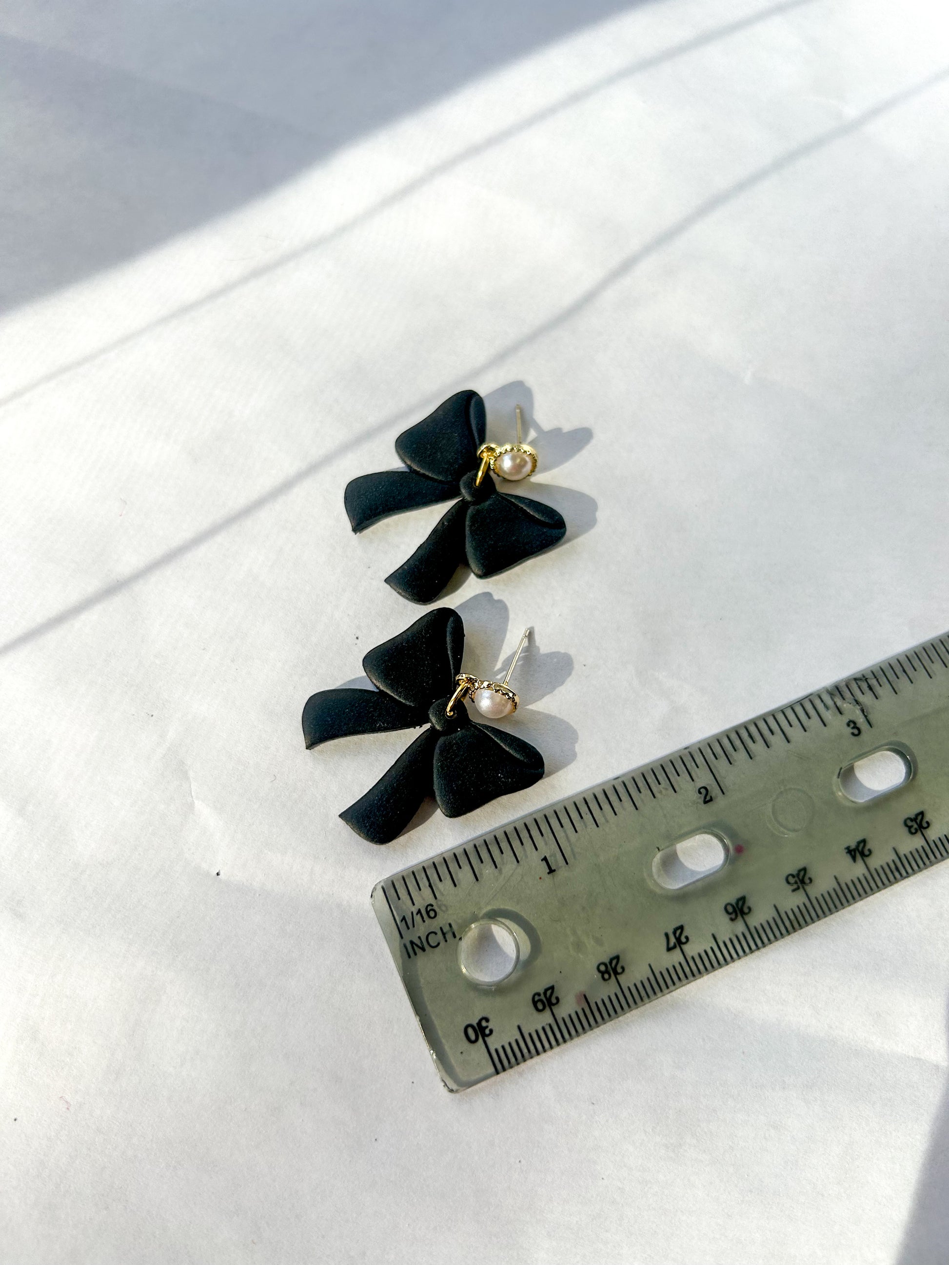 Black bow-shaped earrings with a pearl on a white surface next to a ruler for scale.