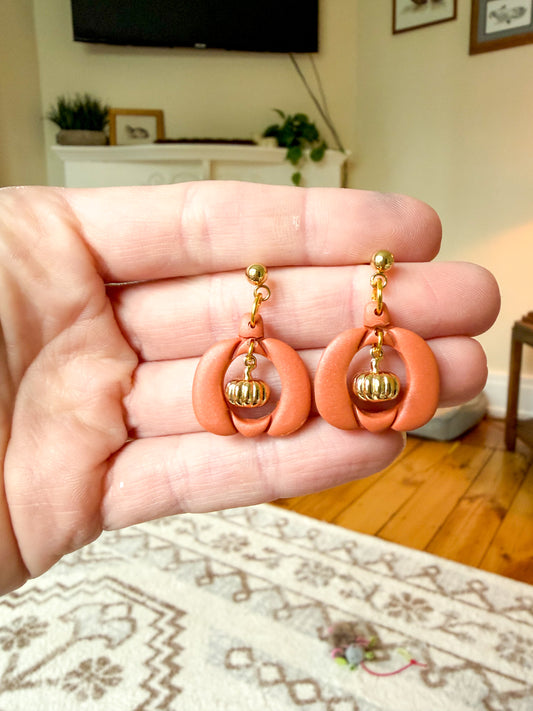 A pair of handmade orange and gold dangle earrings held in a person's hand, with a blurred living room background.