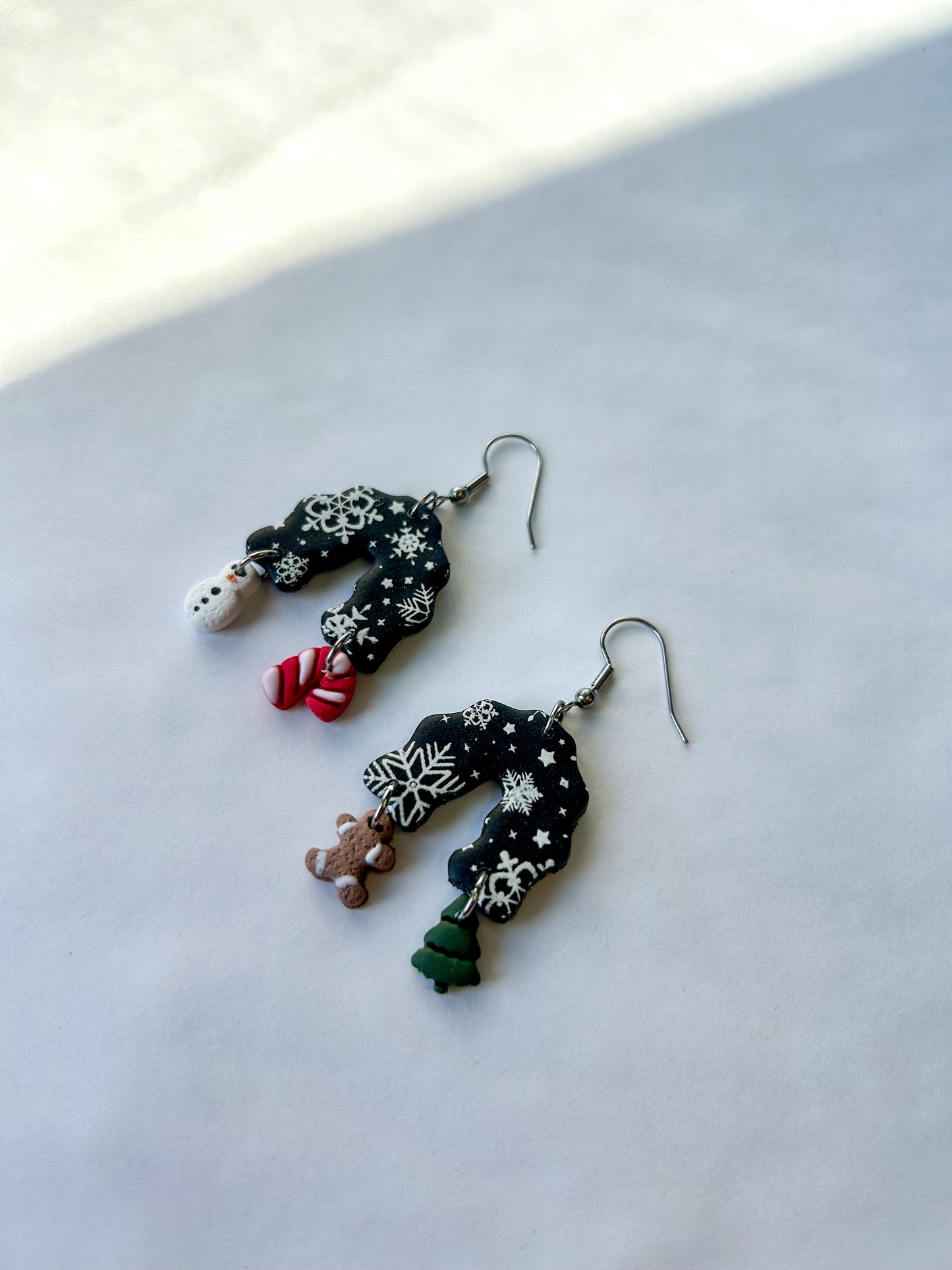 Pair of black earrings on a light background. Black arch dangles with a snowflake design. The four charm hanging off of the earrings are a tree, a gingerbread man, a candy cane, and a snowman