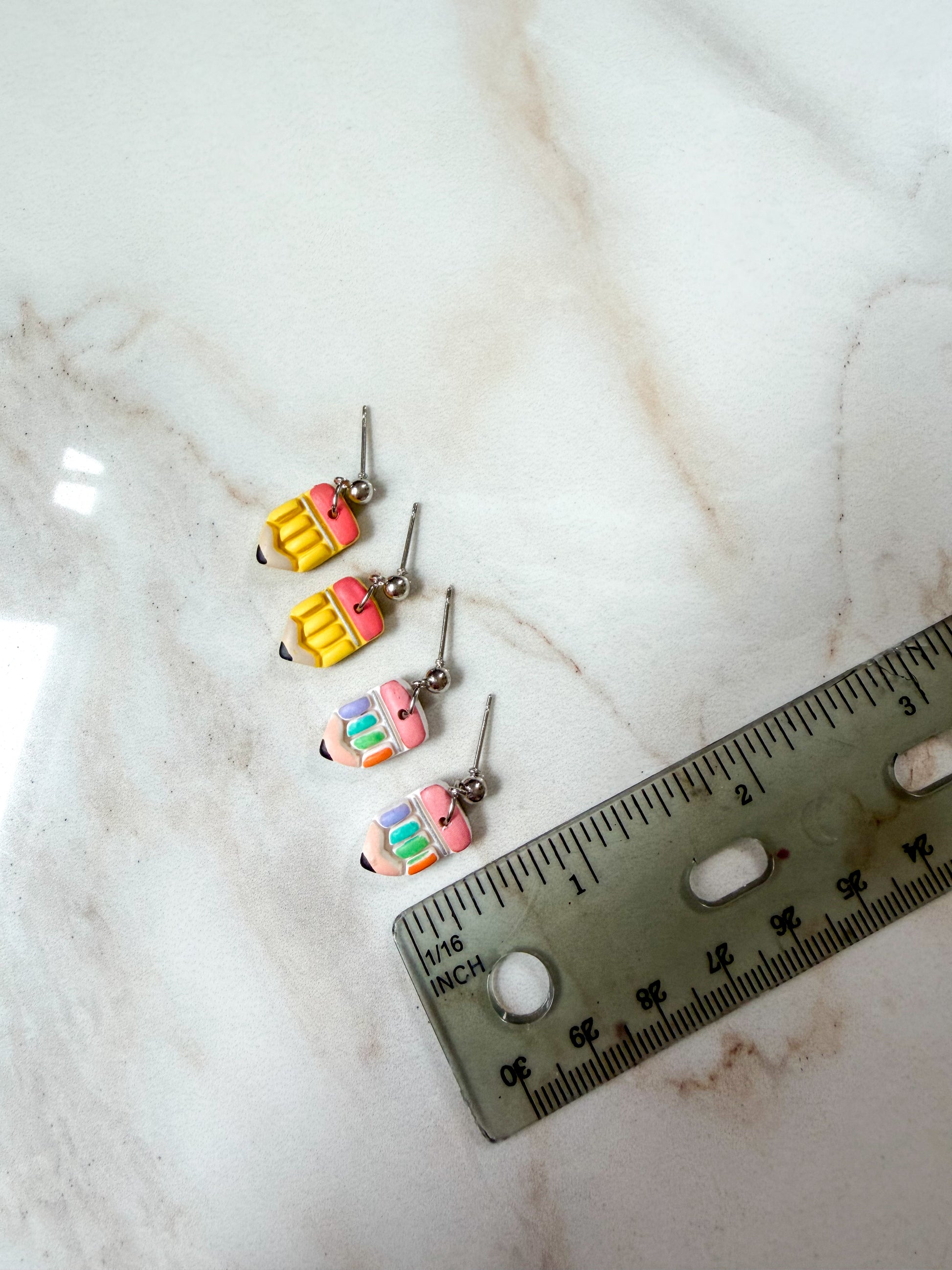 Two pairs of colorful pencil-shaped dangle earrings arranged next to a ruler on a white marble surface. About 3/4 inches long. 