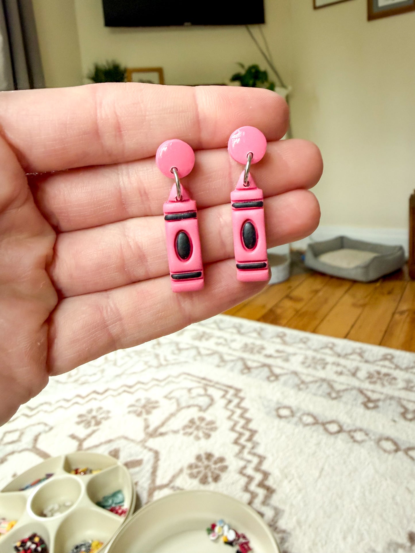 A hand holds a pair of pink polymer clay earrings shaped like crayons, with black and silver details.
