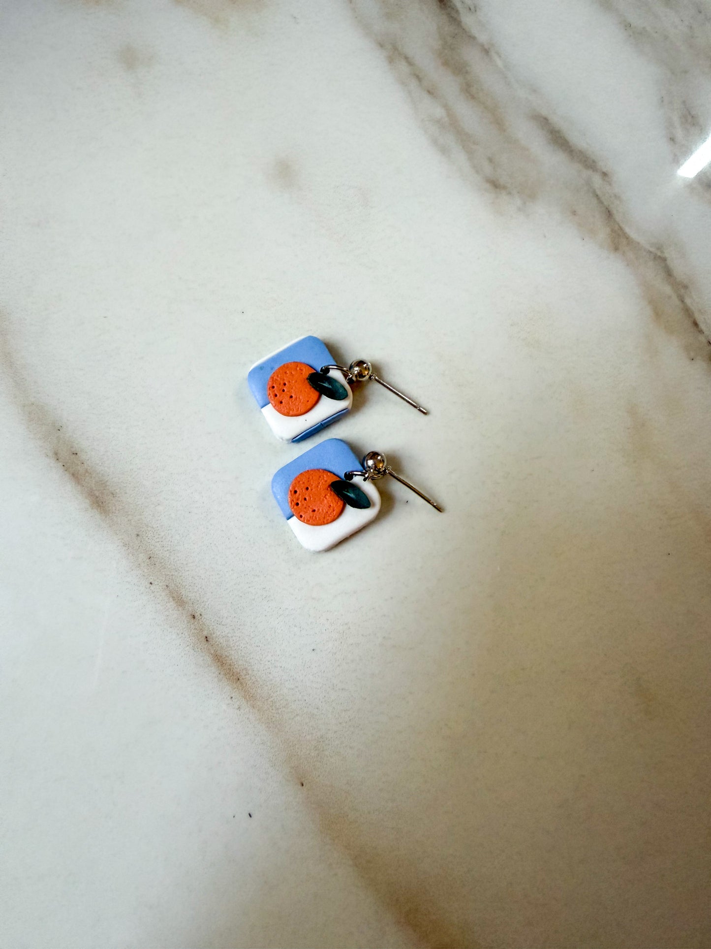 A pair of handmade square dangle earrings with a blue and white background, featuring a central orange circle and a small green leaf, resting on a light marble surface.