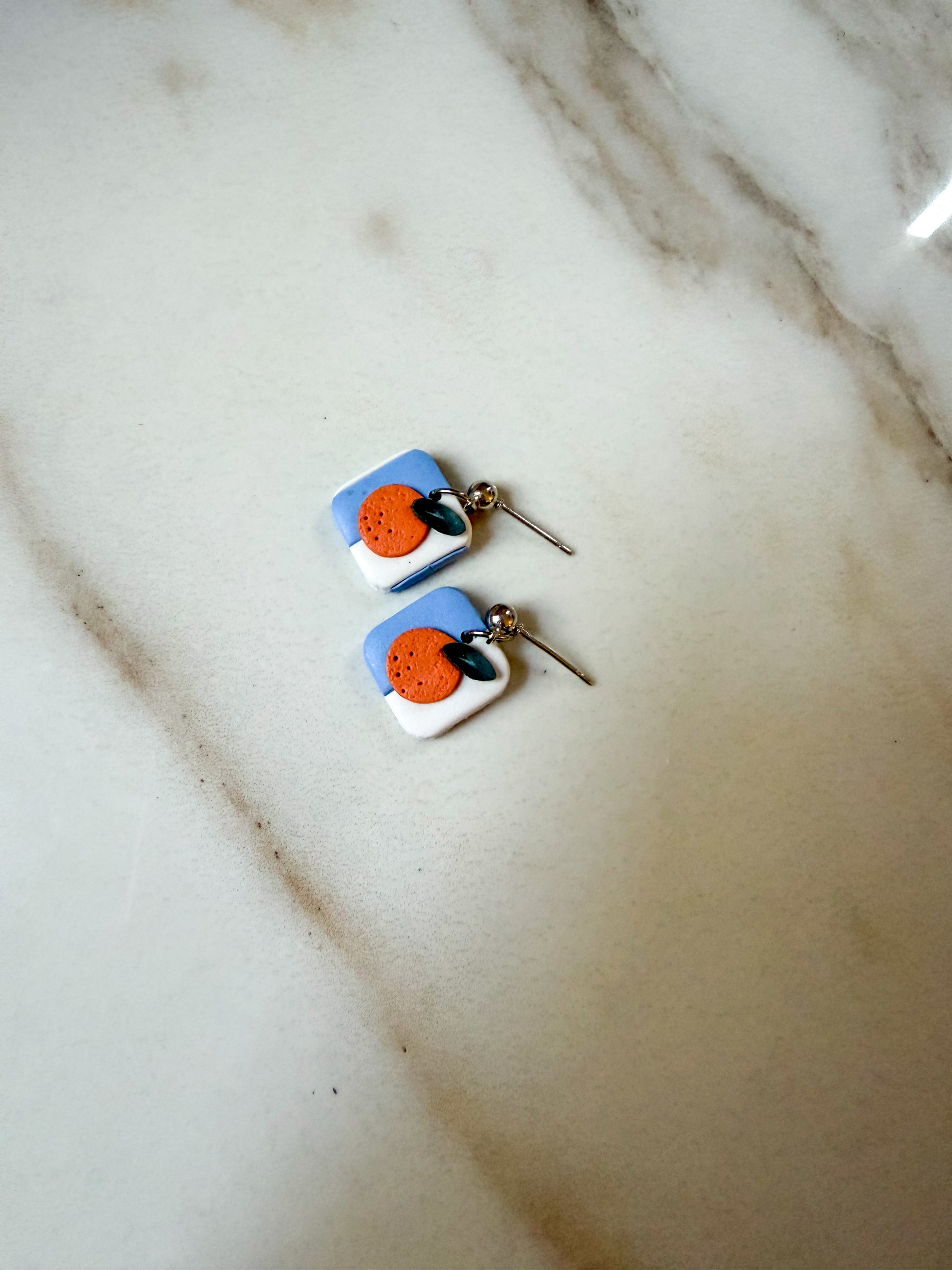 A pair of handmade square dangle earrings with a blue and white background, featuring a central orange circle and a small green leaf, resting on a light marble surface.