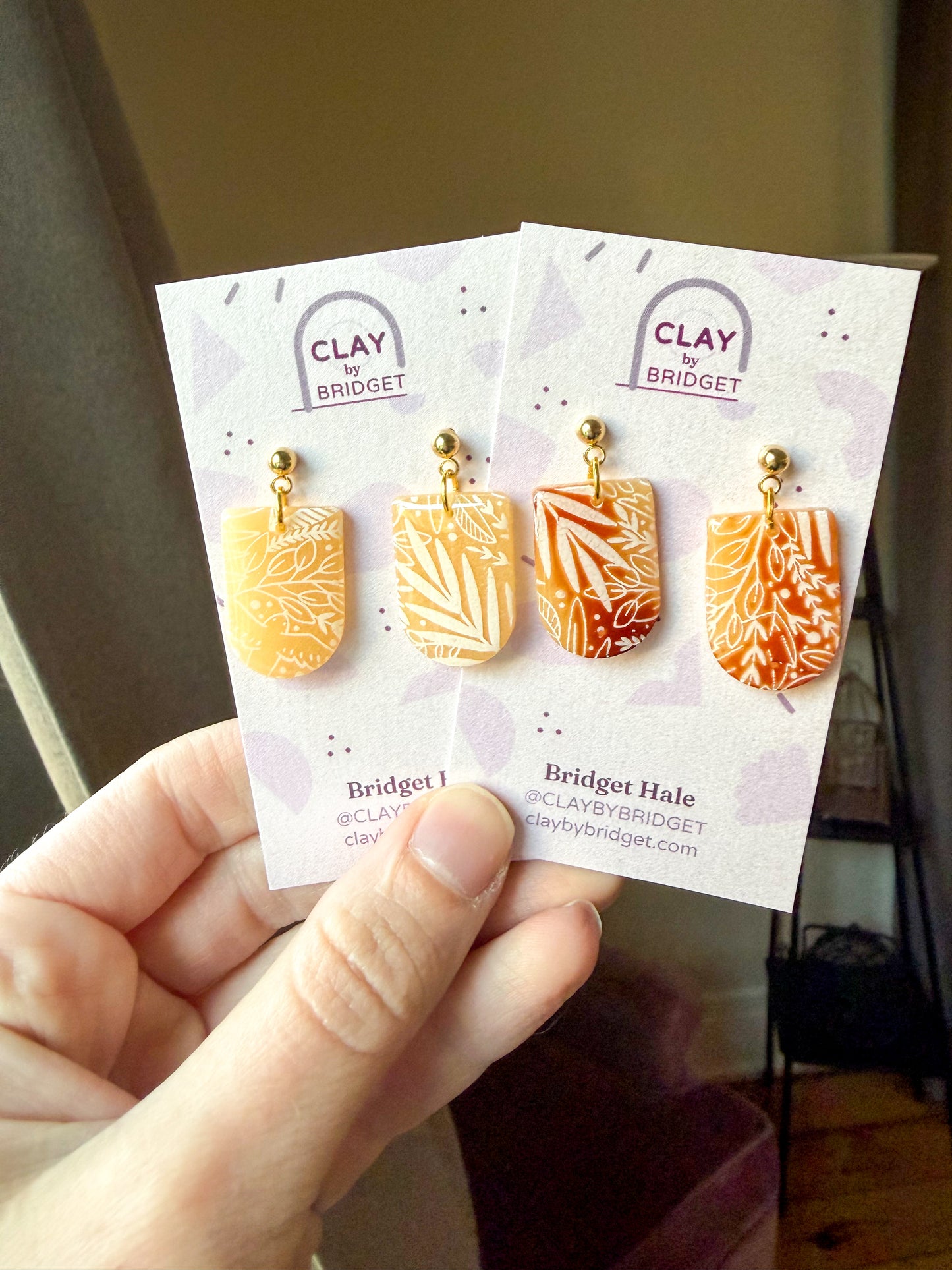 Hands holding two pairs of earrings on purple earring cards. They are transluscent with white floral painted designs. 