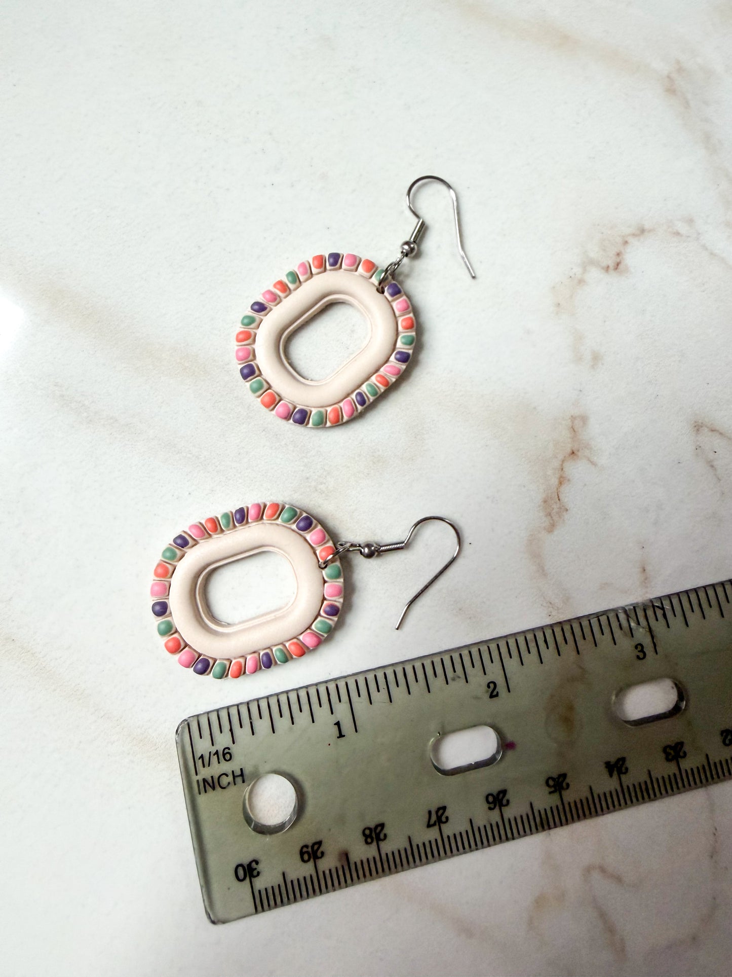 Pair of earrings with colorful painted detailing on a marble surface with a ruler for scale.