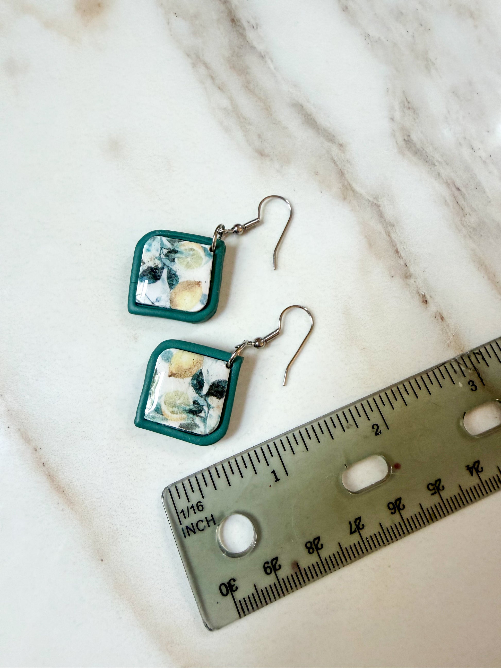 A pair of handmade square dangle earrings with teal green borders and a white and lemon center, displayed on a white marble surface next to a metal ruler for scale. About 2 inches long. 
