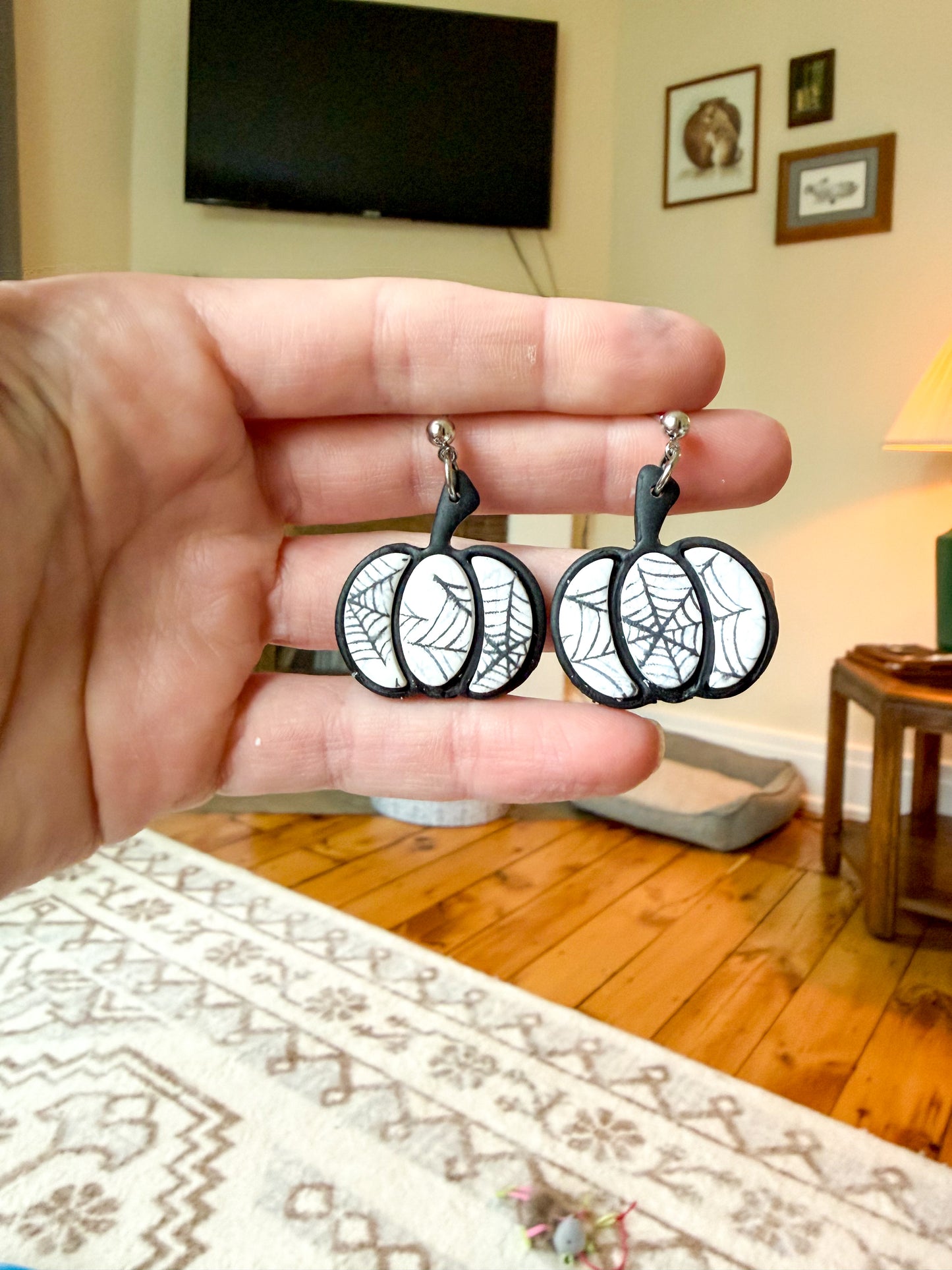 A hand holds a pair of black and white spiderweb pumpkin earrings in front of a living room background with a TV, lamp, and wooden floor.