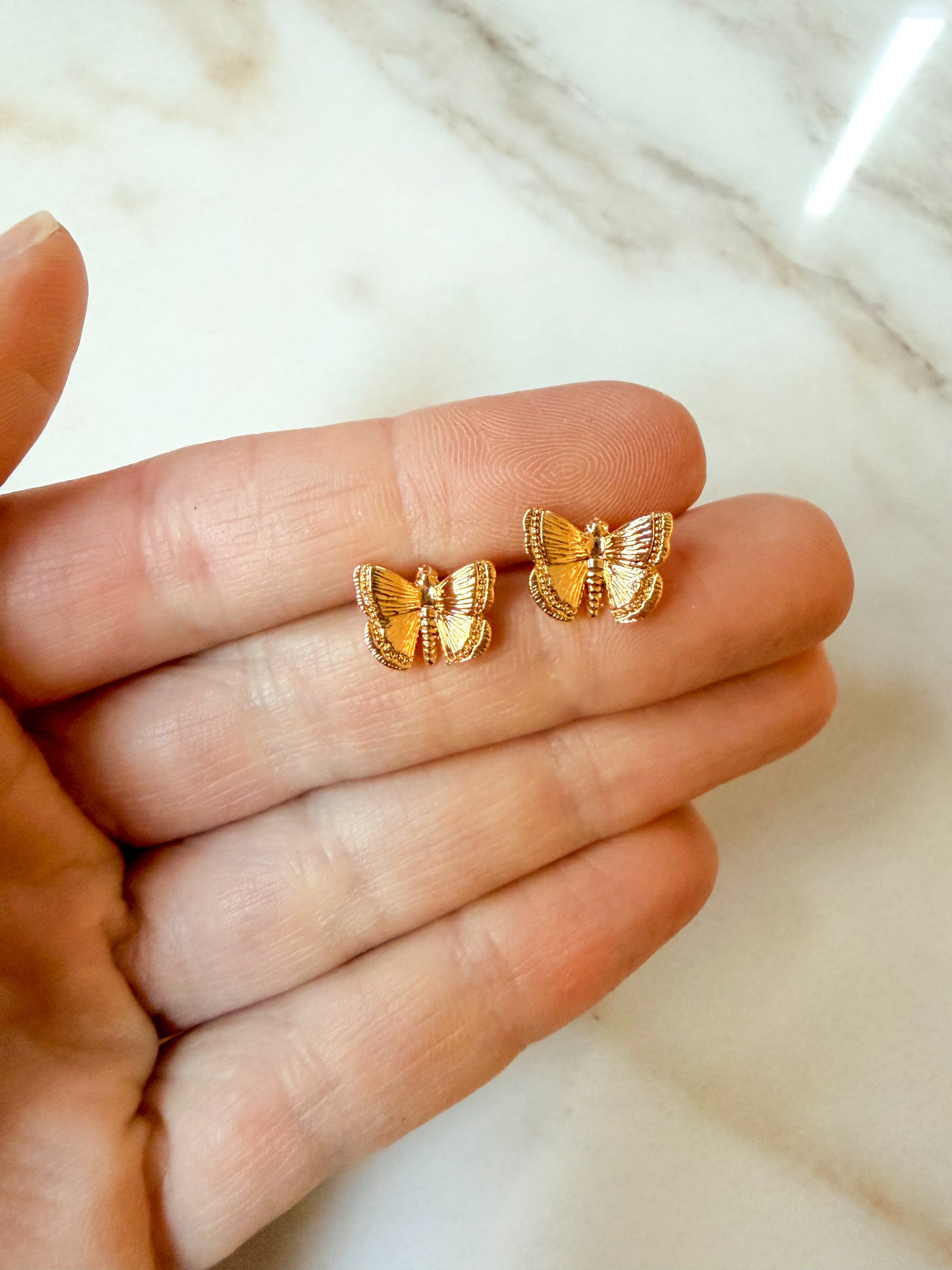 A pair of small gold butterfly stud earrings resting on an open hand, with a light marble surface in the background.