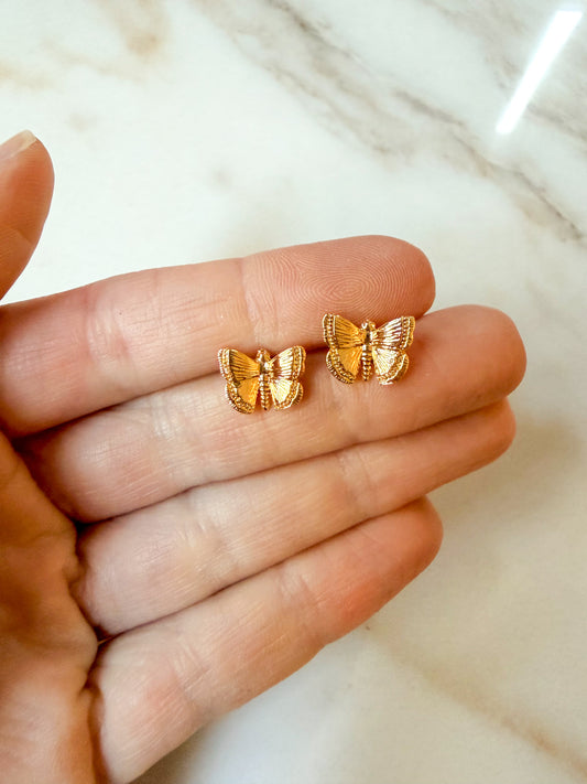 A pair of small gold butterfly stud earrings resting on an open hand, with a light marble surface in the background.