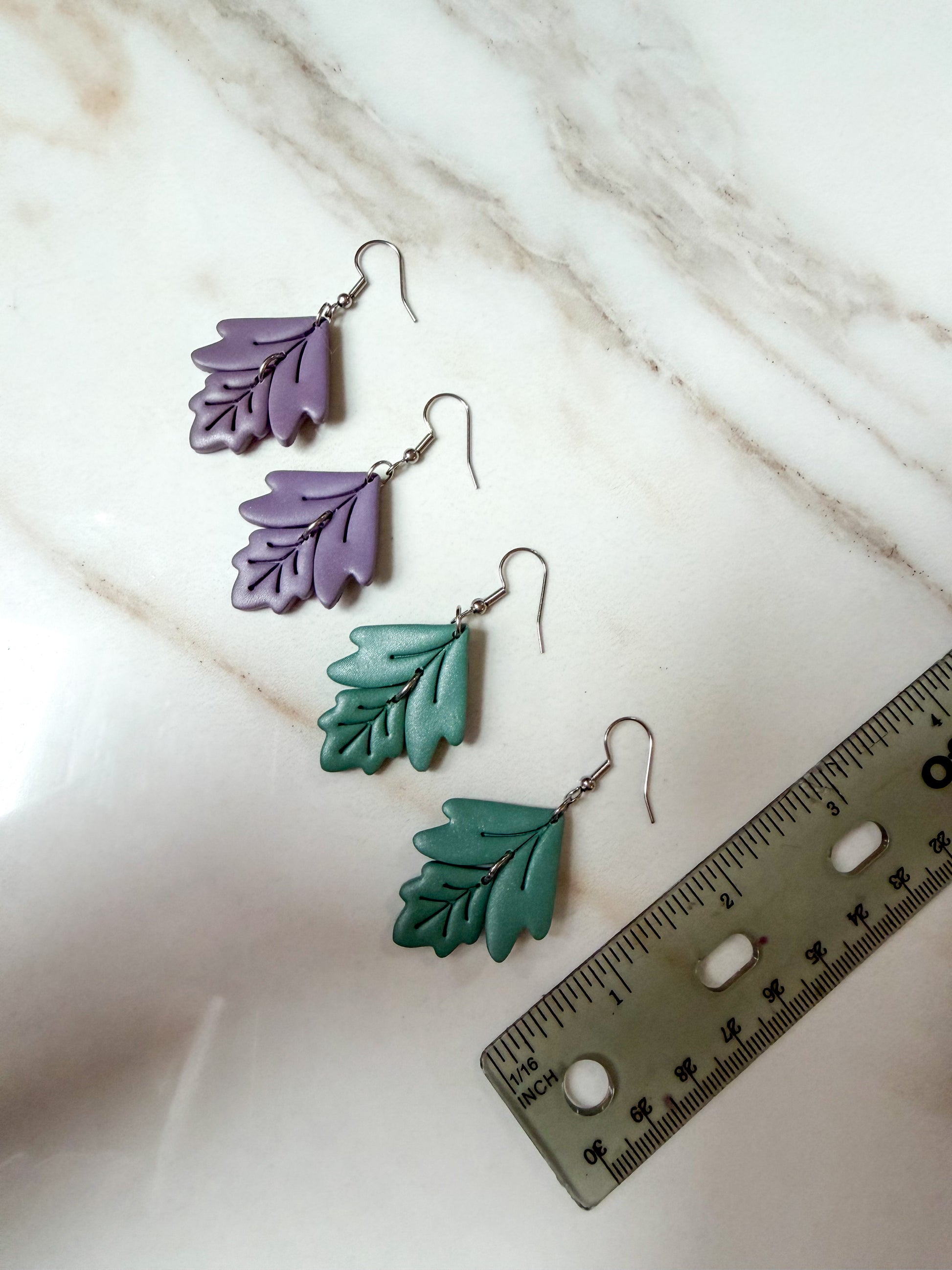Four leaf-shaped drop earrings in alternating lavender purple and teal green, displayed on a white marble surface next to a ruler for scale. Around 2 inches in length. 